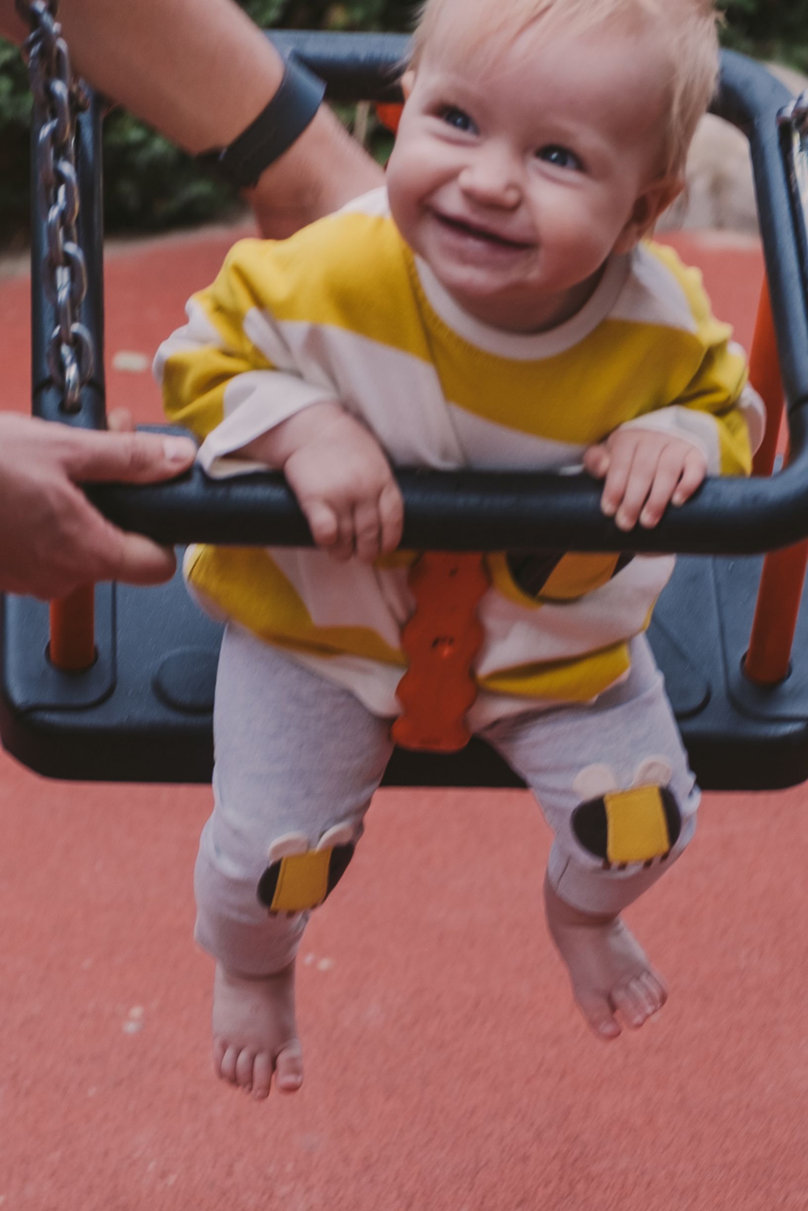 A baby wearing a yellow shirt and gray pants plays on playground equipment with a joyful expression.