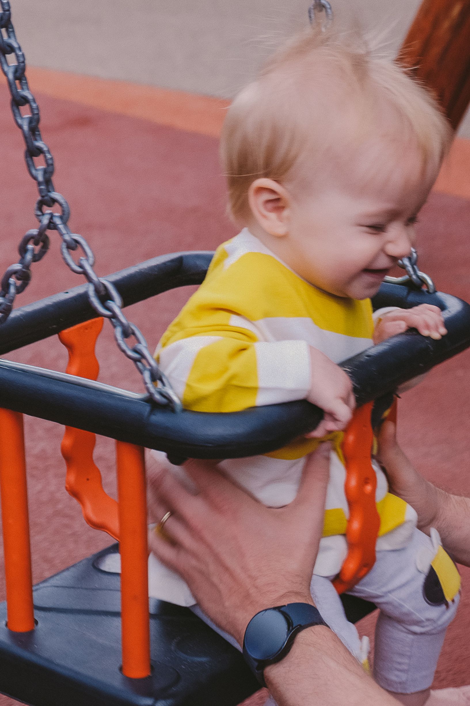 A young child in a yellow shirt being helped on a playground swing set with orange support poles.