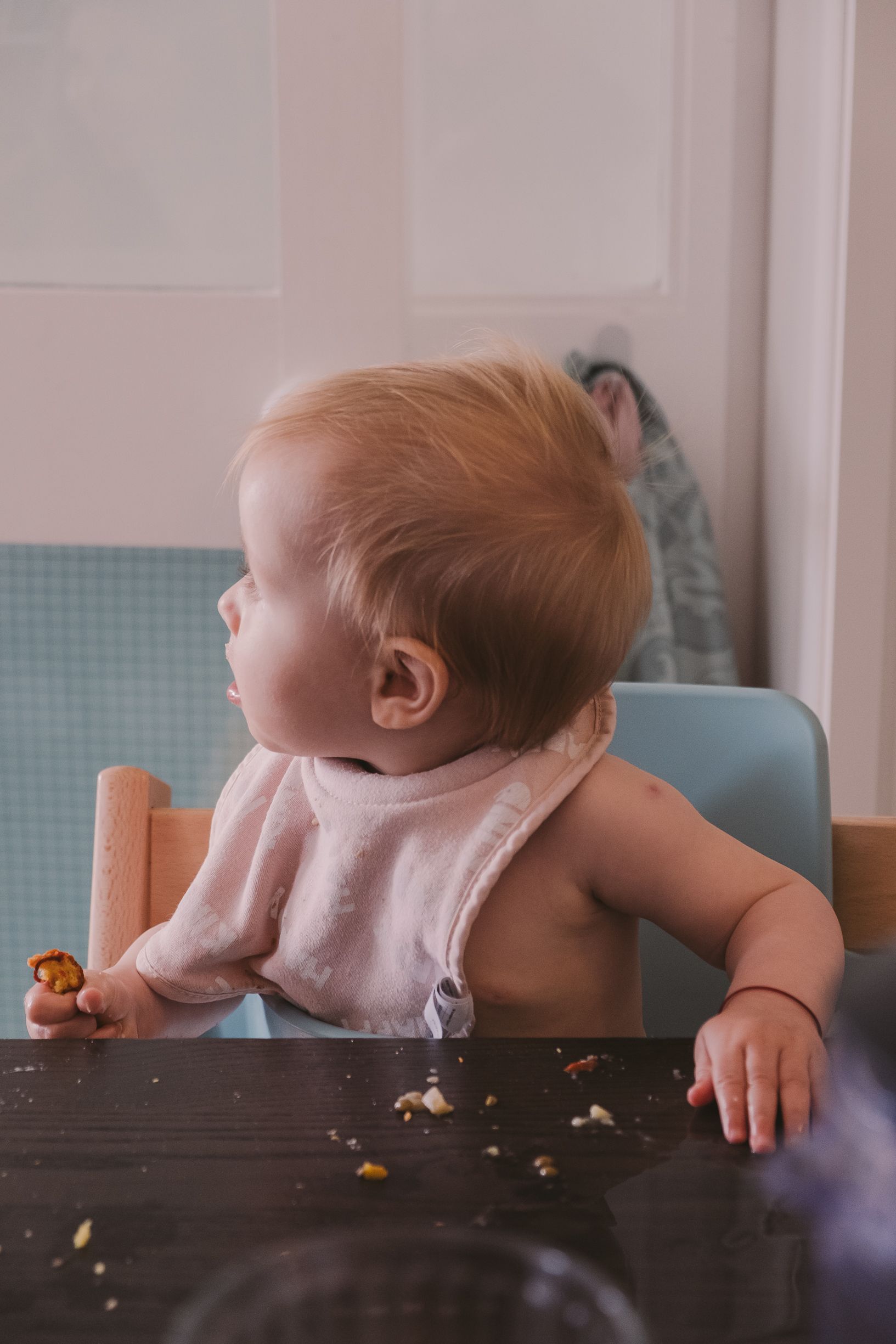 A baby leans over a dark wooden table surface reaching for scattered cookie crumbs.