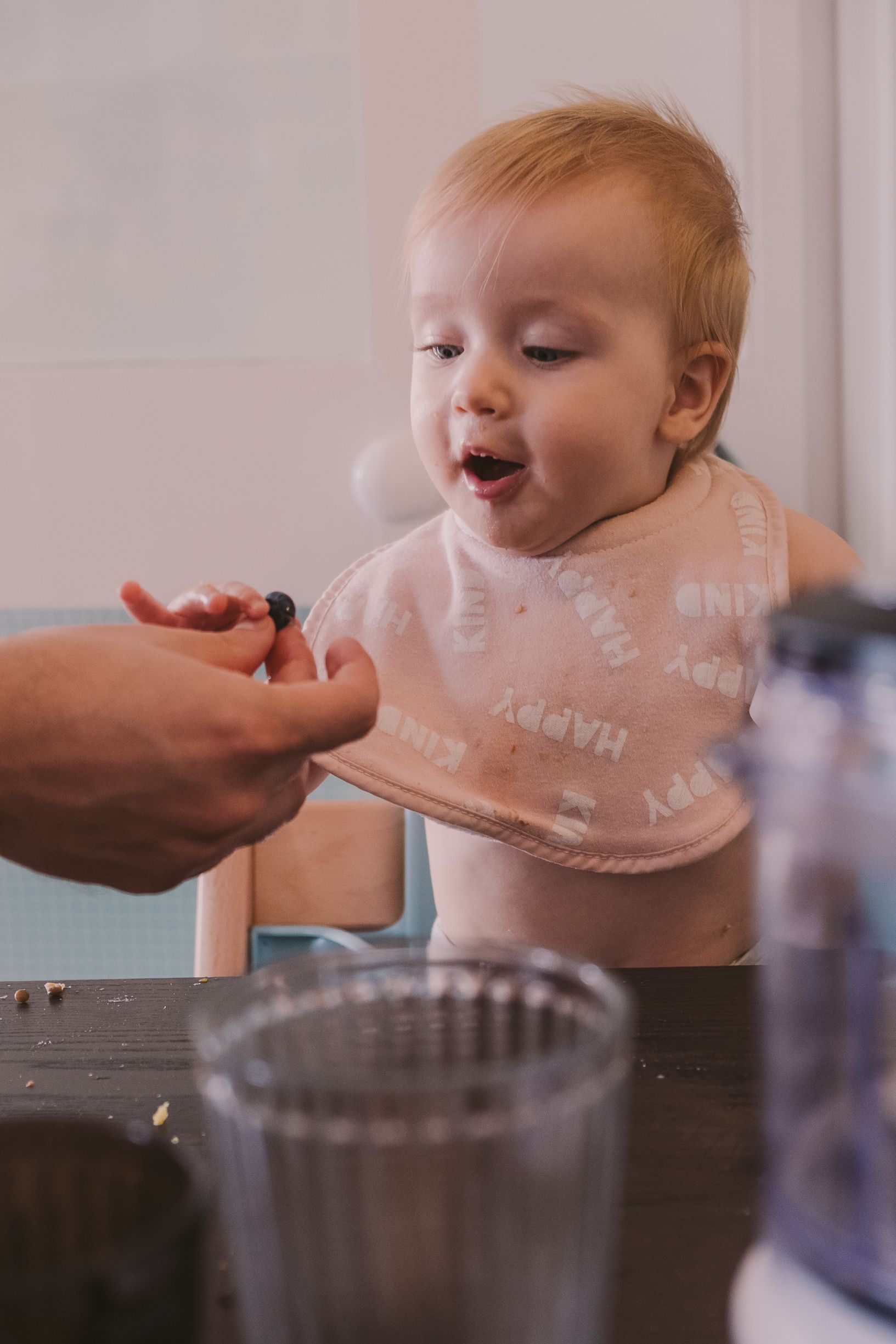 A baby wearing a light pink bib opens their mouth while being fed at a table with glasses.