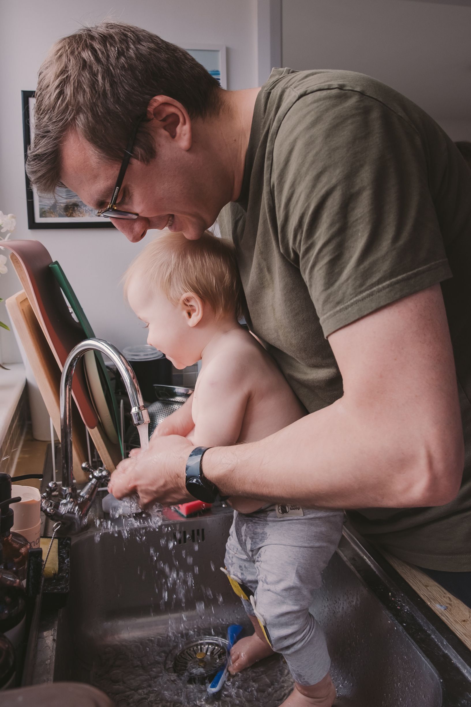 A parent and small child wash dishes together at the kitchen sink, with water splashing as they work.