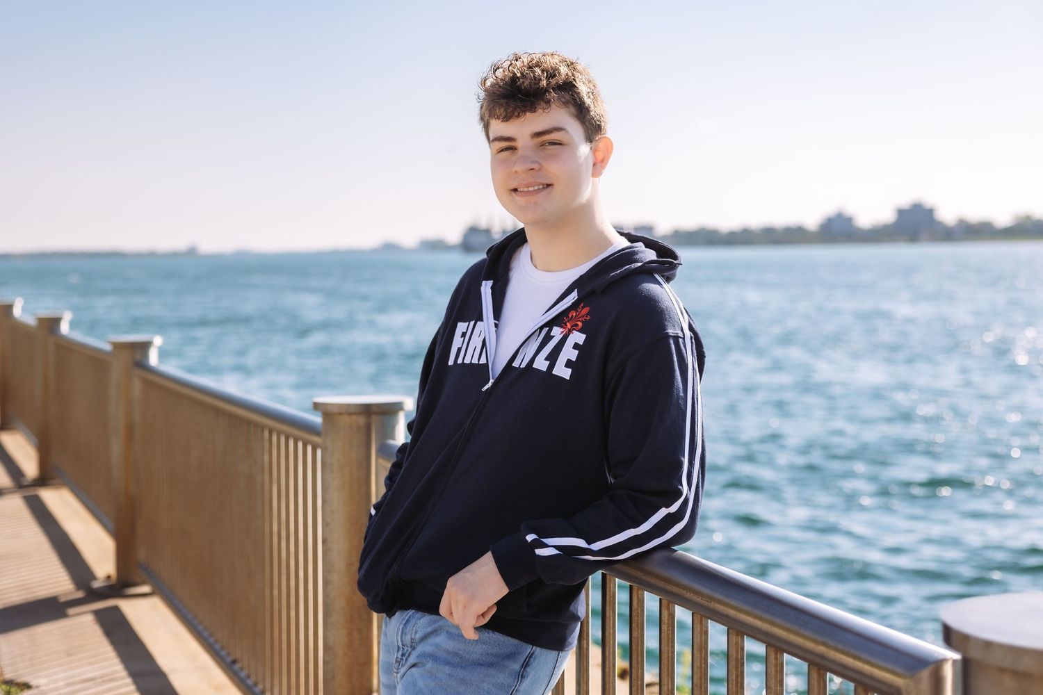 A person in a Nike hoodie stands on a wooden deck overlooking a scenic waterfront view on a sunny day.
