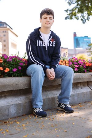 A person in a navy hoodie and jeans sits on a concrete ledge with colorful flowers and fallen leaves in the background.
