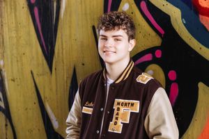 A performer in a brown jacket stands in front of a yellow wall with colorful decorative elements.
