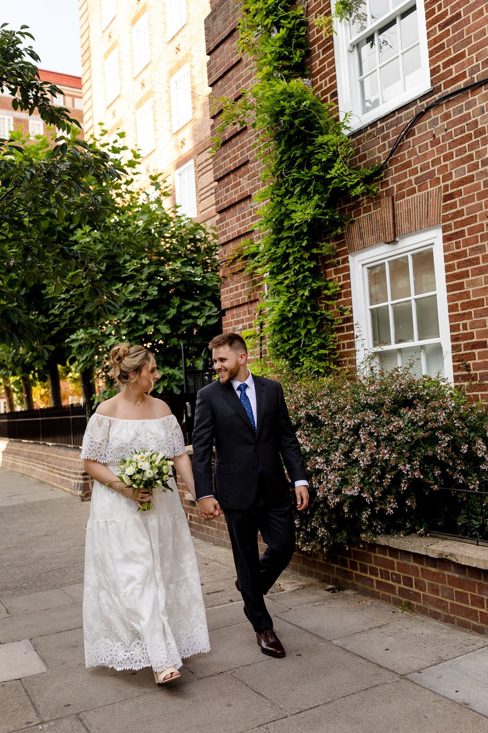 A couple walking together on a sidewalk by a brick building with ivy, the bride in a lace dress holding a bouquet.