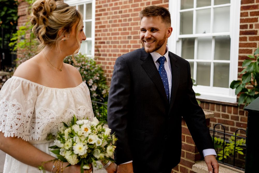 A romantic wedding moment captured outside a brick building with white windows and lush greenery.