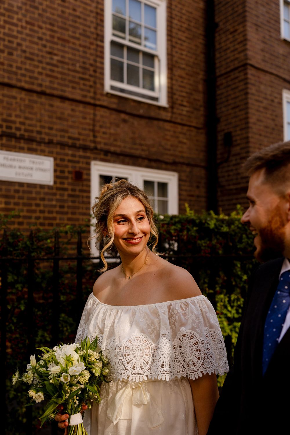 A bride in an off-shoulder lace wedding dress smiles while holding a white and green bouquet against a brick building.
