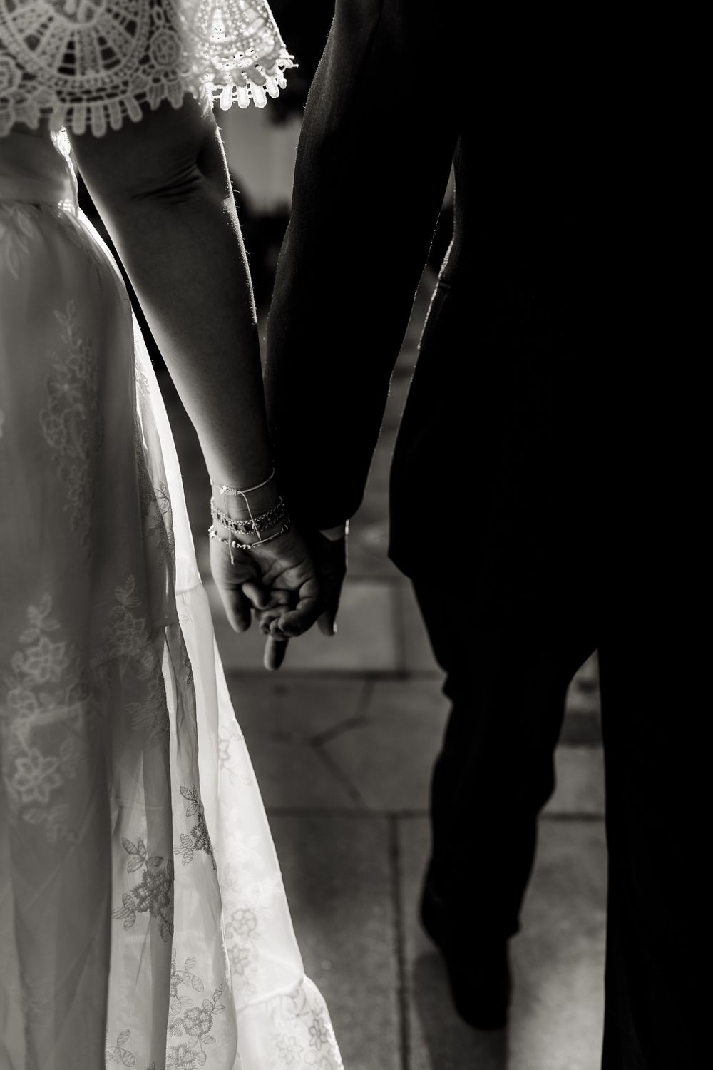 Black and white photo showing a bride's white lace wedding dress hem next to groom's dark pants and shoes.
