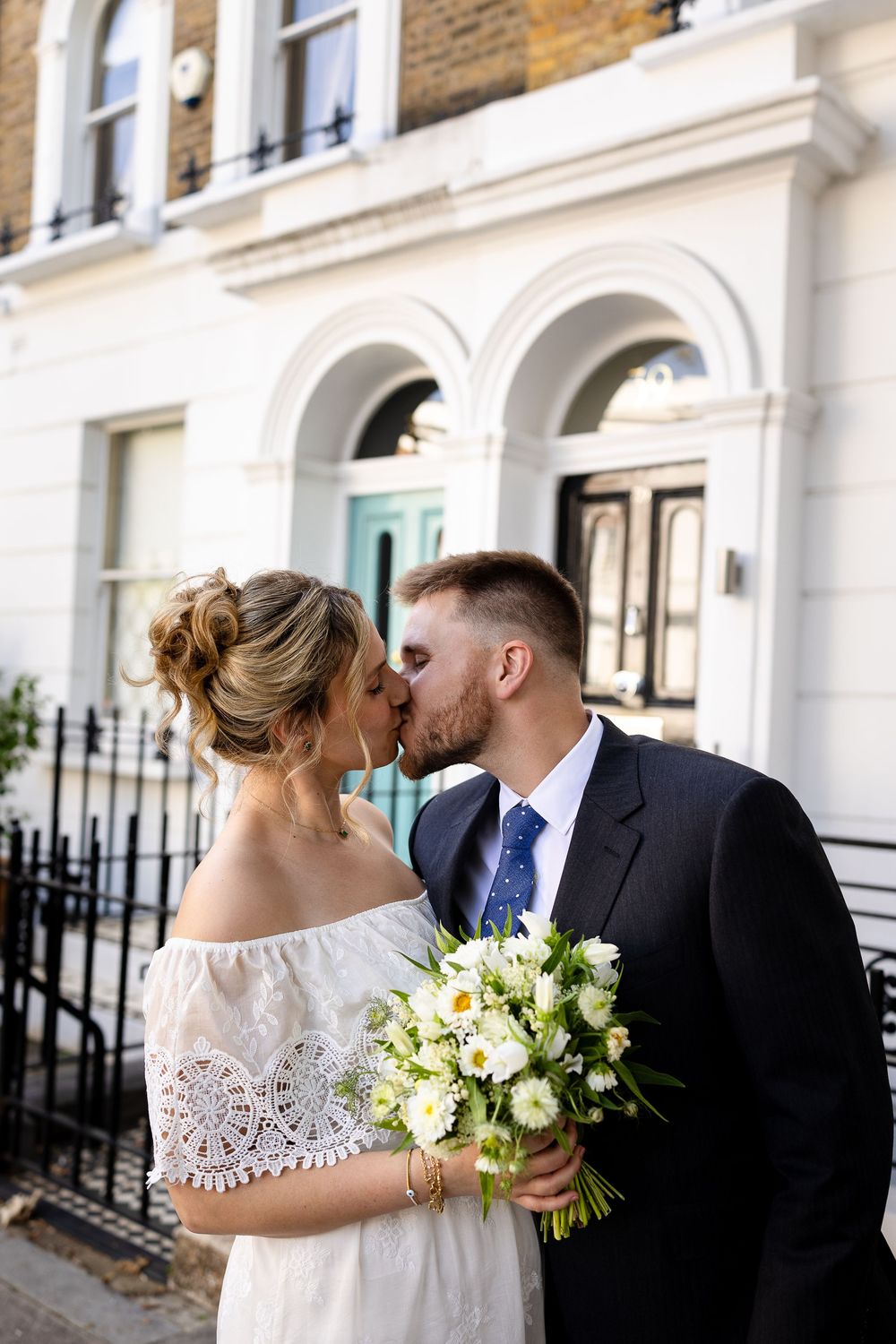 A romantic moment between a couple on their wedding day outside a white townhouse with arched windows.