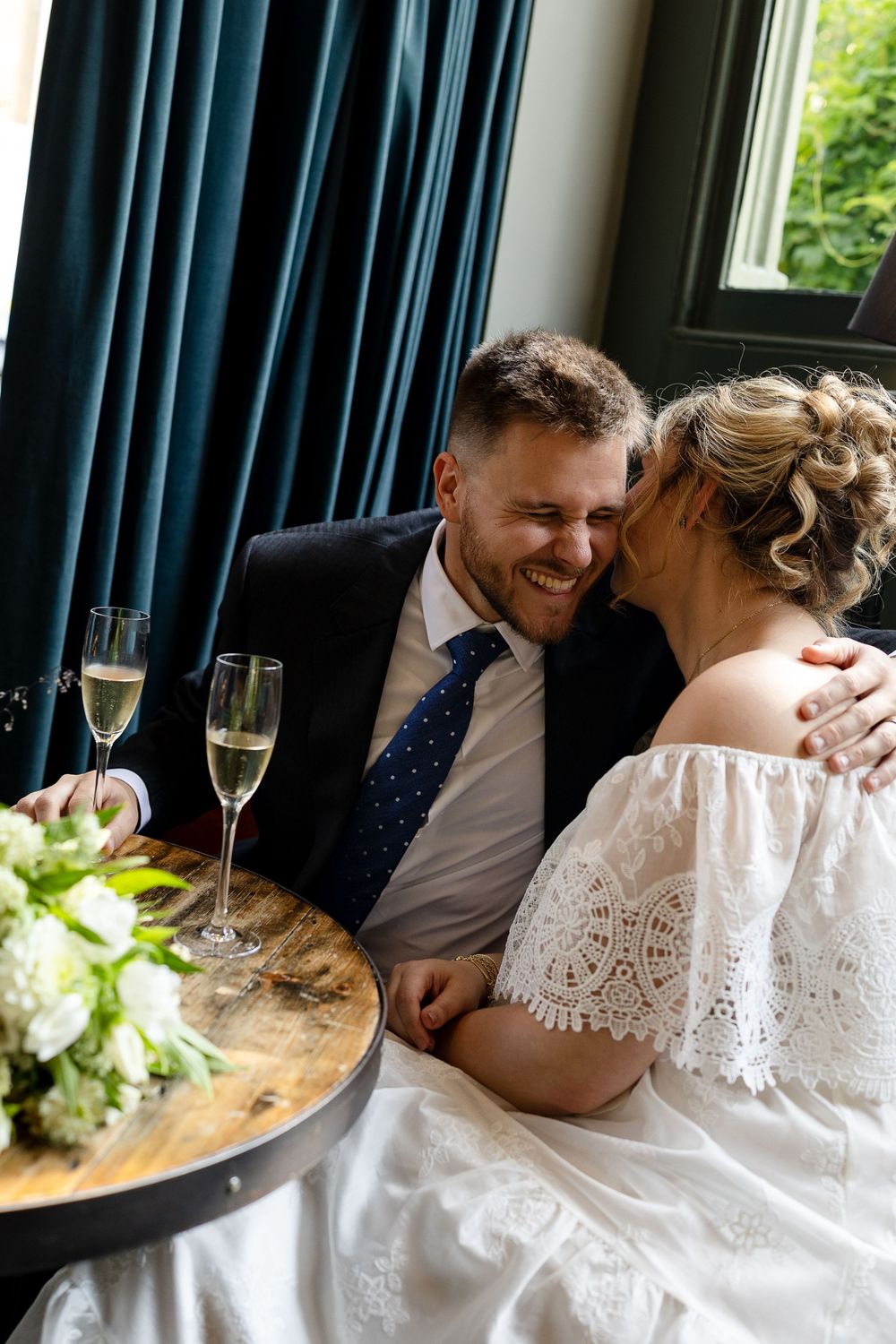A joyful wedding moment captured near a window with champagne and flowers on a rustic table.