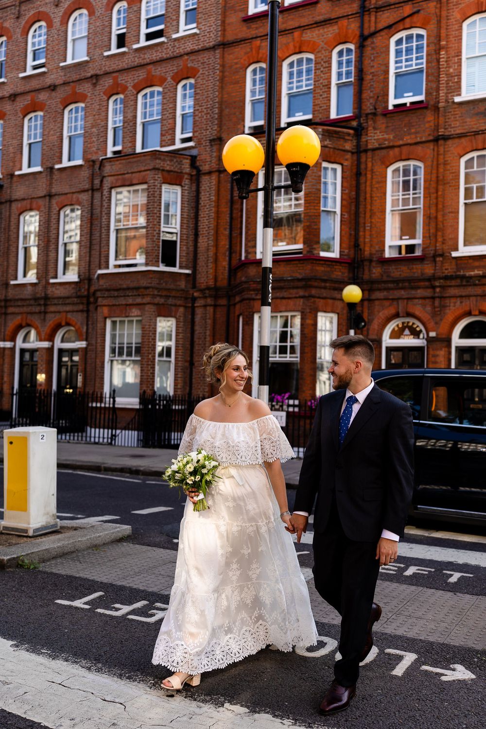 A bride in a white lace dress and groom in a dark suit walk together on a brick-lined street near historic buildings.