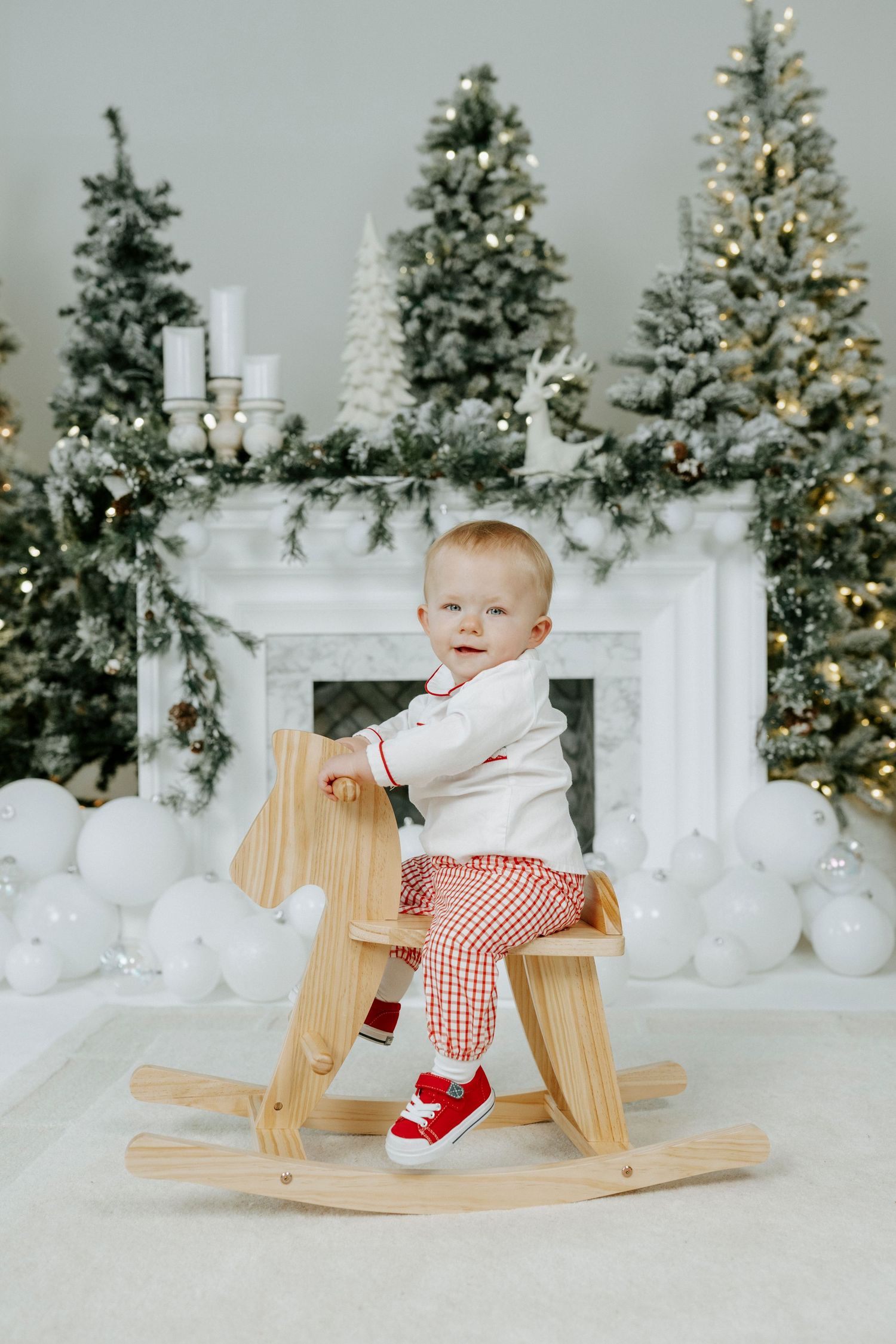 A wooden rocking horse toy photographed in front of decorated Christmas trees and white holiday decor.