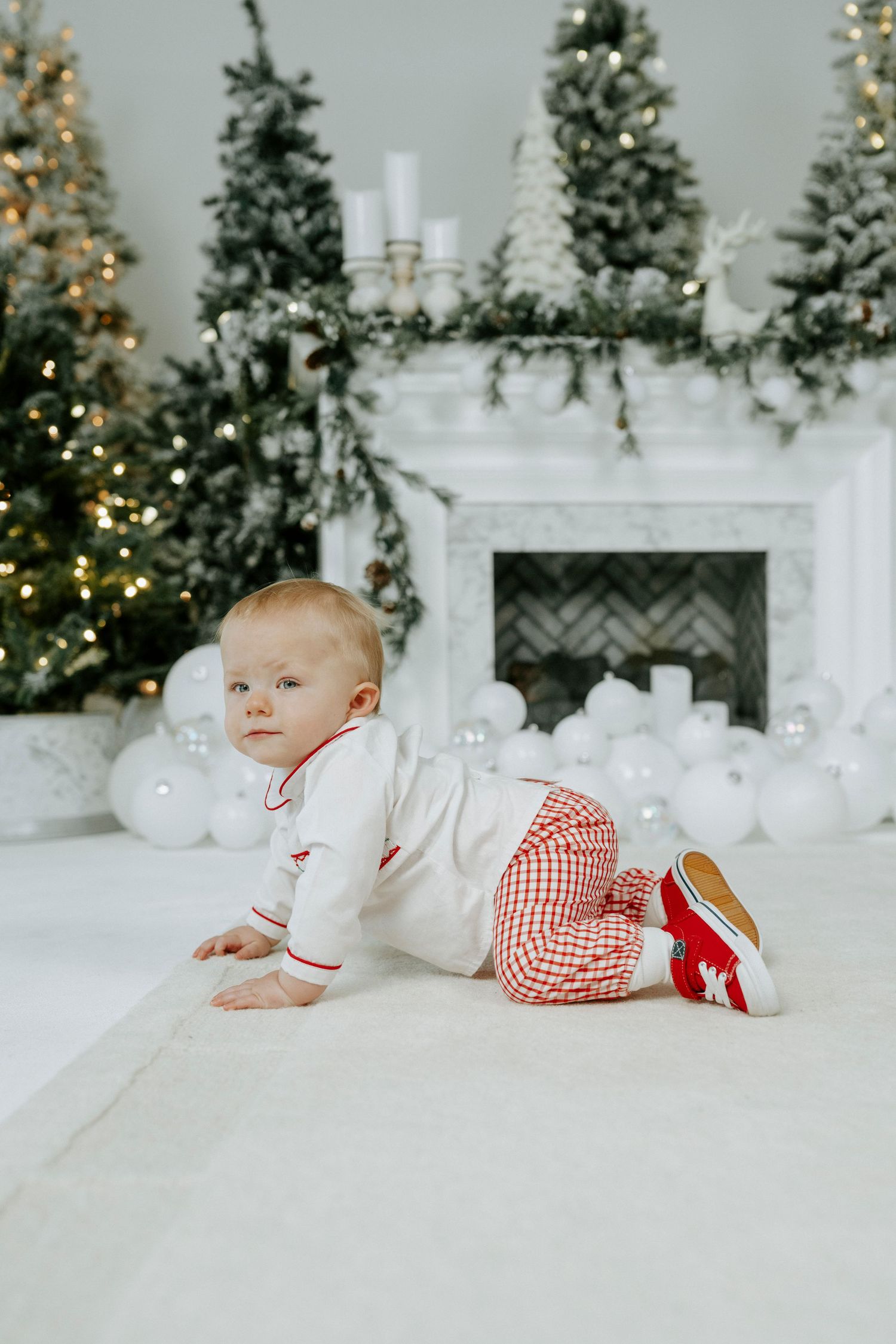 A festive holiday scene with a baby in red and white clothing laying on a white blanket near decorated Christmas trees.