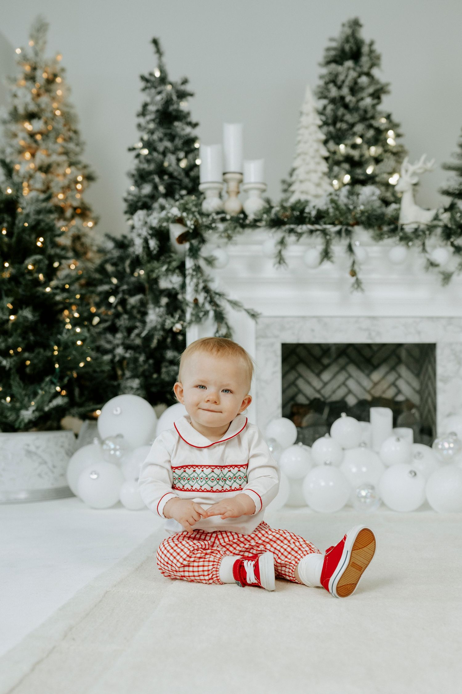 Festive holiday scene with decorated Christmas trees, white balloons, and a baby in red and white outfit sitting on floor.