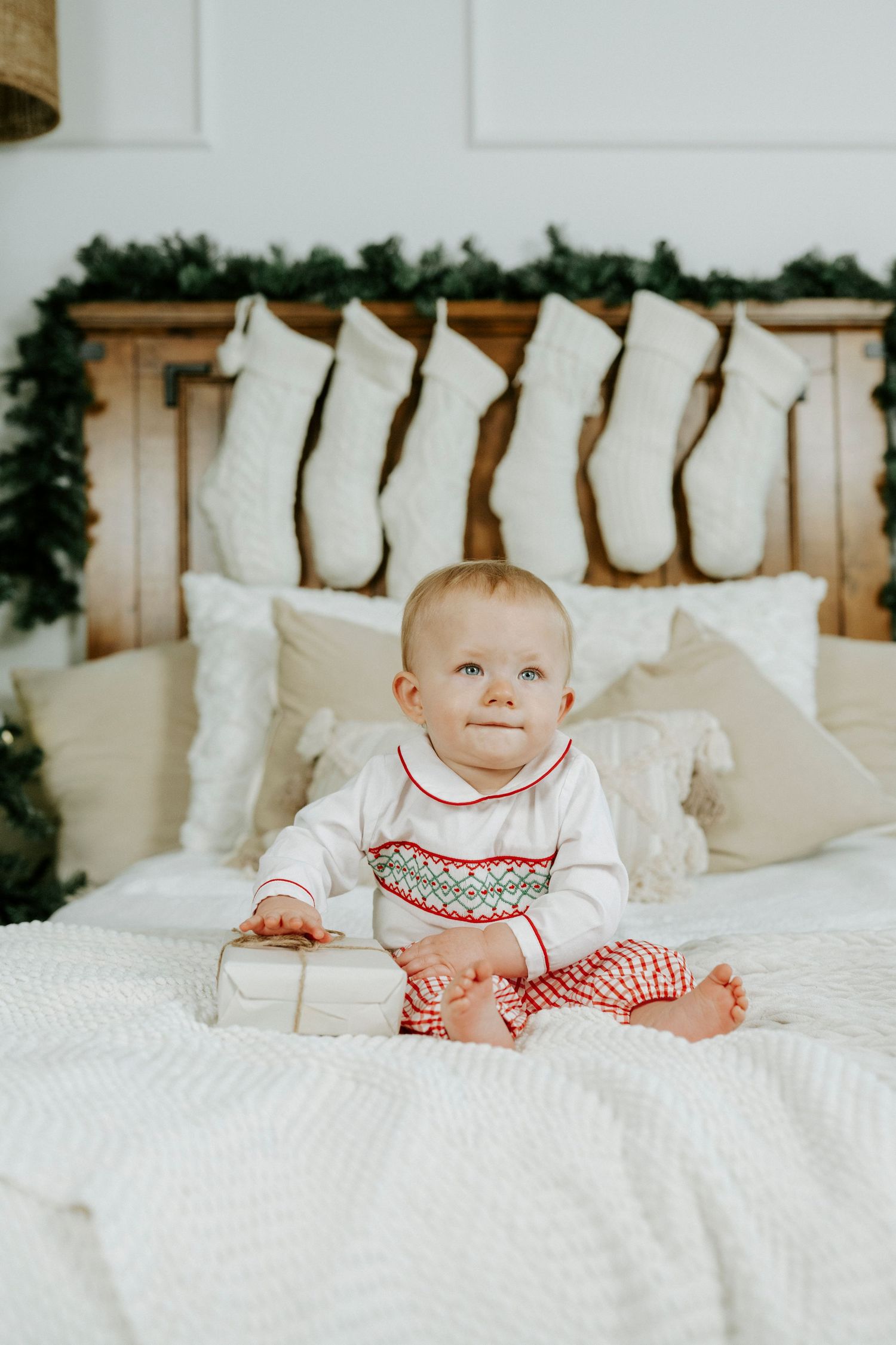 Festive Christmas scene with white stockings hanging on headboard and baby sitting on white bedding in holiday attire.