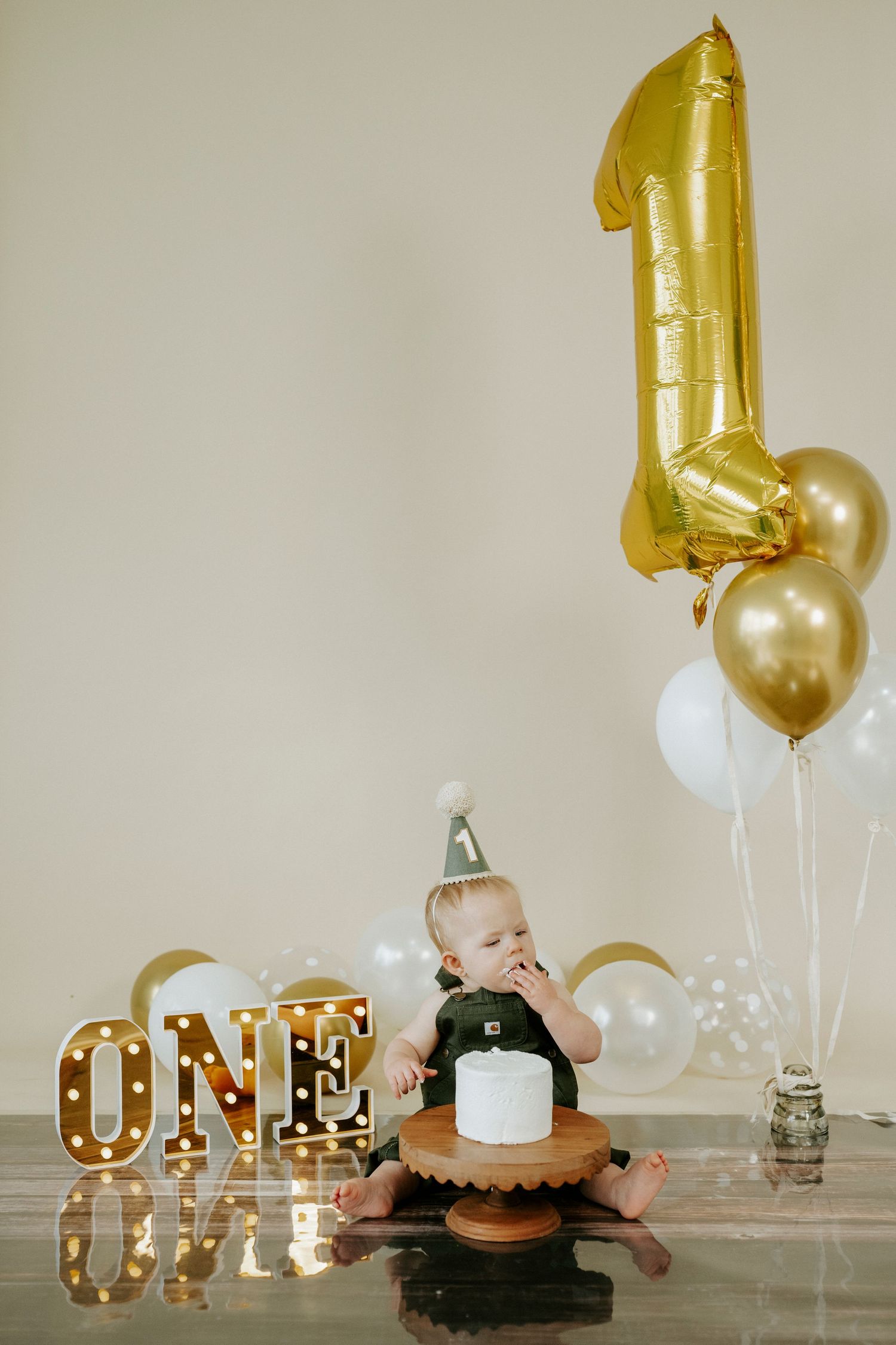 First birthday celebration setup with gold marquee letters, balloons, and a cake on reflective surface.