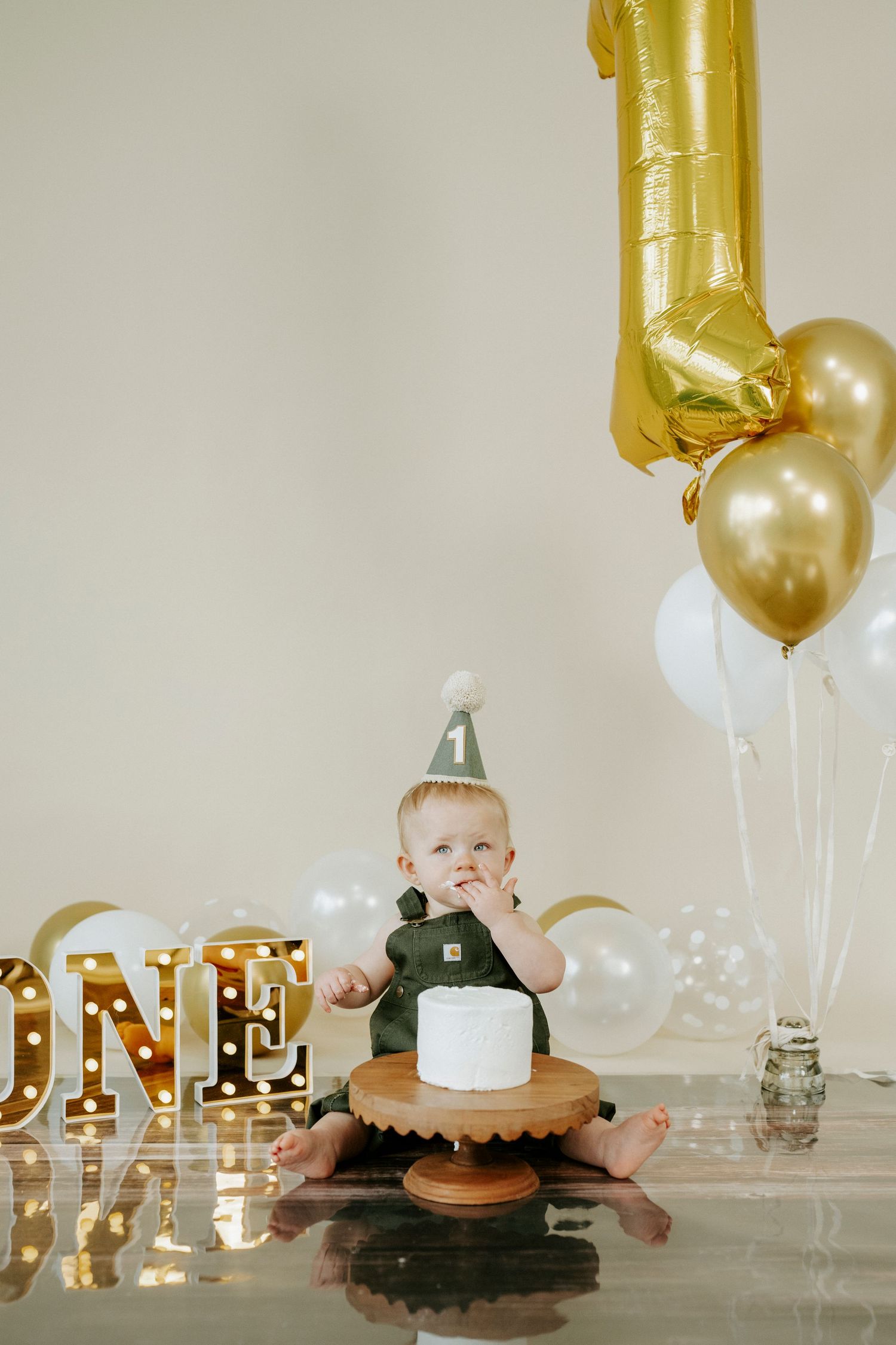 First birthday celebration setup with gold and white balloons, illuminated ONE sign, and small cake on wooden stand.