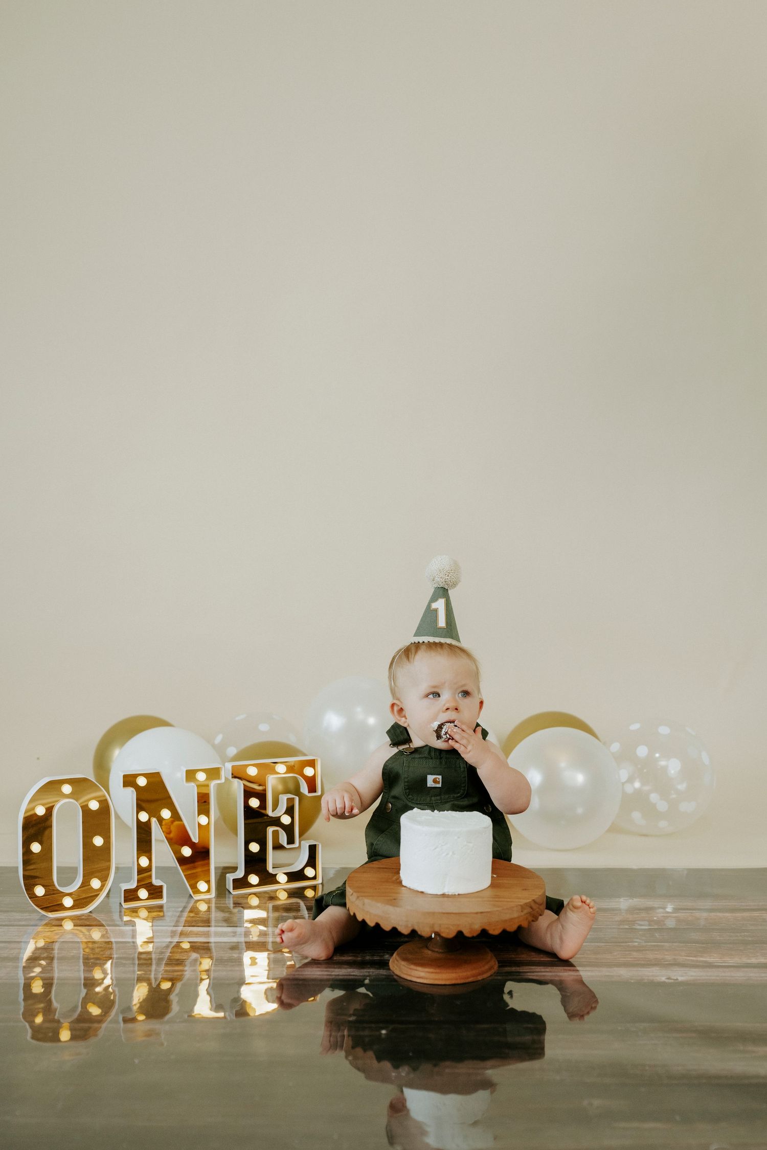 Baby in party hat sits with smash cake beside illuminated 'ONE' marquee letters and balloons for first birthday celebration.