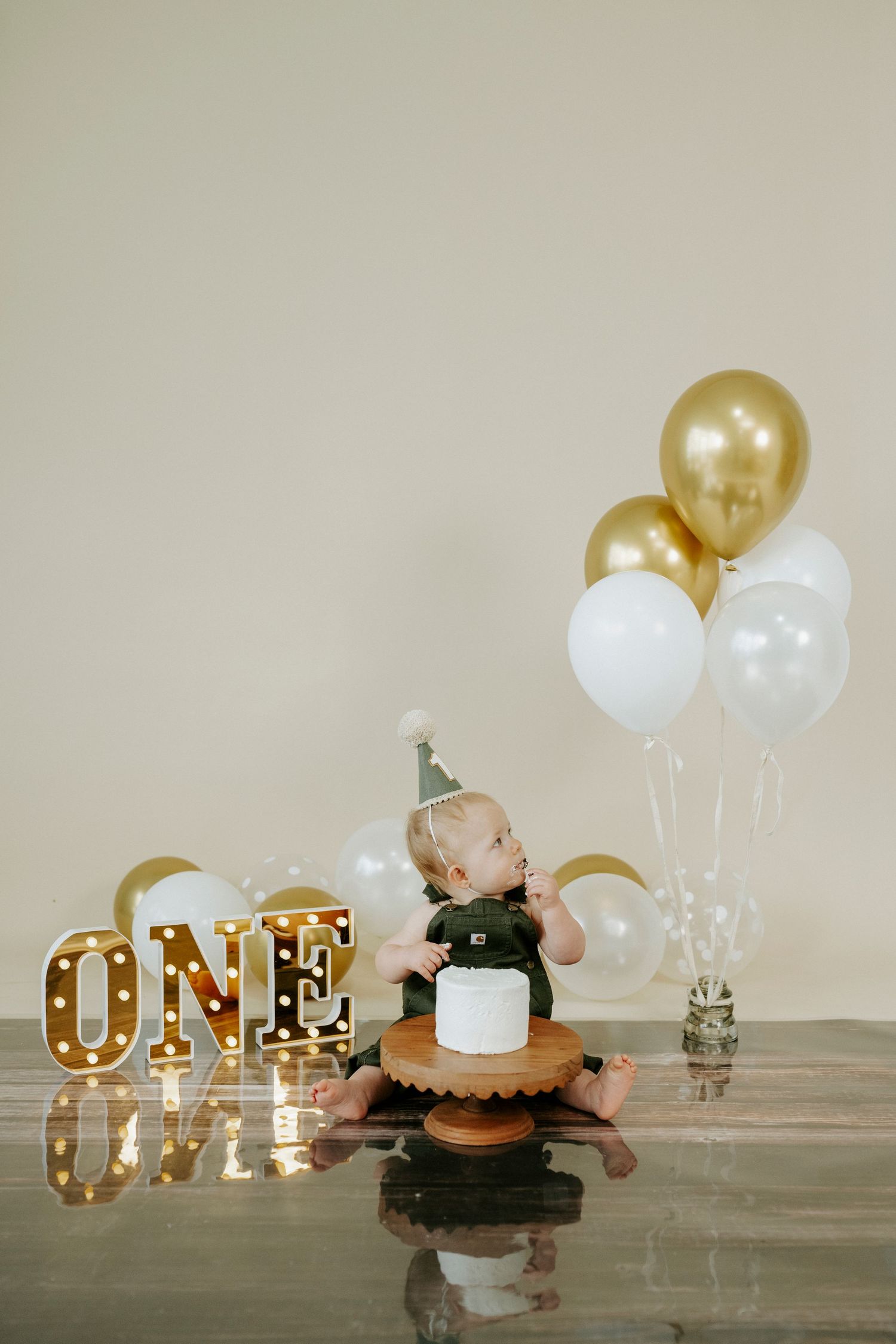 Elegant first birthday setup featuring gold and white balloons with illuminated ONE sign on mirrored surface.