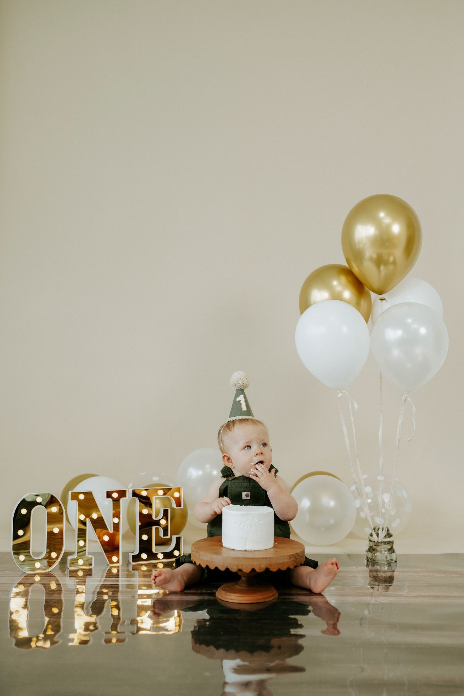 First birthday setup with illuminated letters and white and gold balloons on a neutral background.