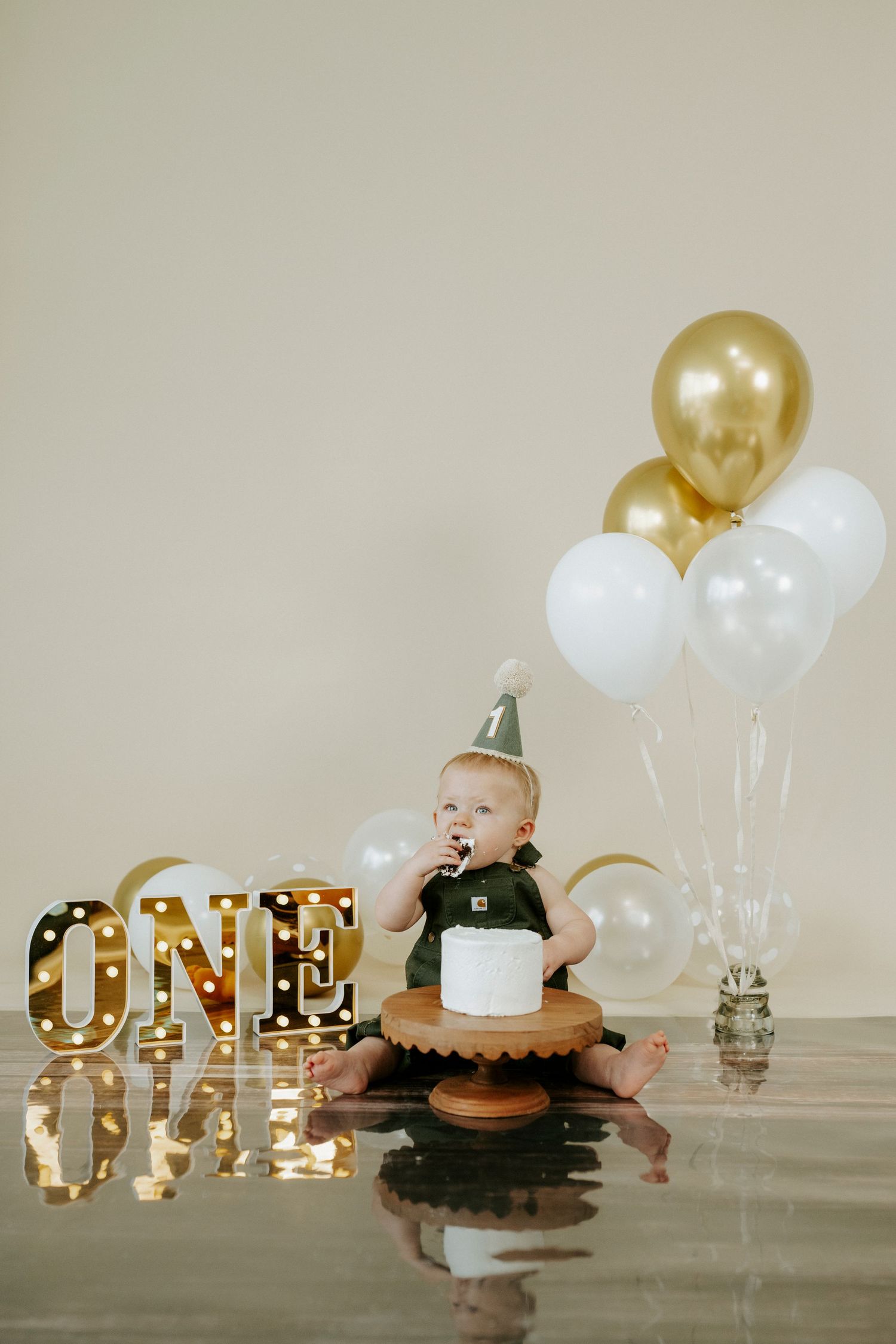 Birthday setup featuring gold marquee ONE sign, white and gold balloons, and cake on wooden pedestal.