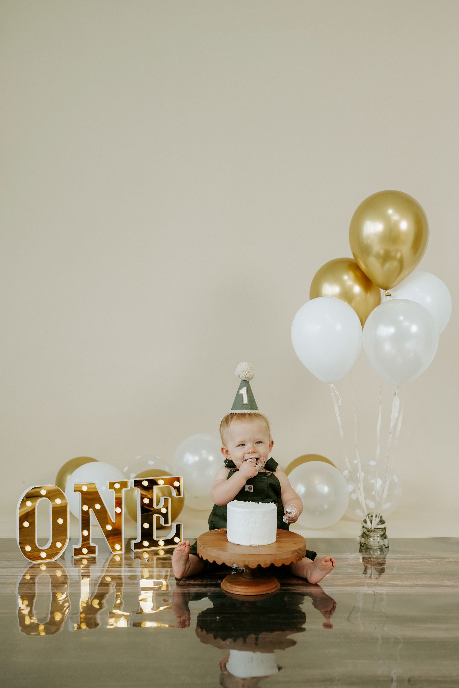 First birthday celebration setup with gold light-up 'ONE' sign and white and gold balloons on wooden surface.