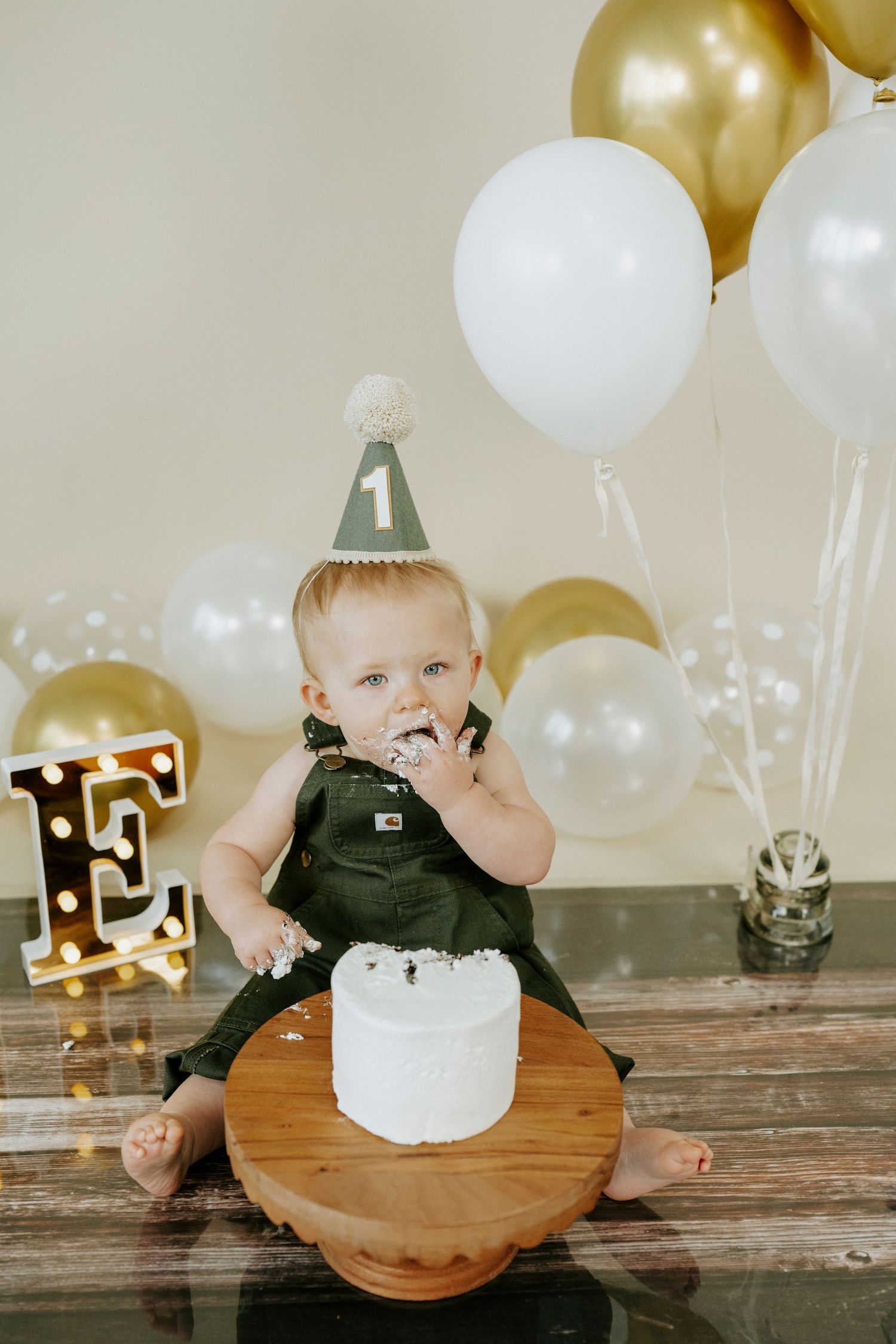 First birthday party scene with gold letter decor, white and gold balloons, and cake on wooden cake stand.