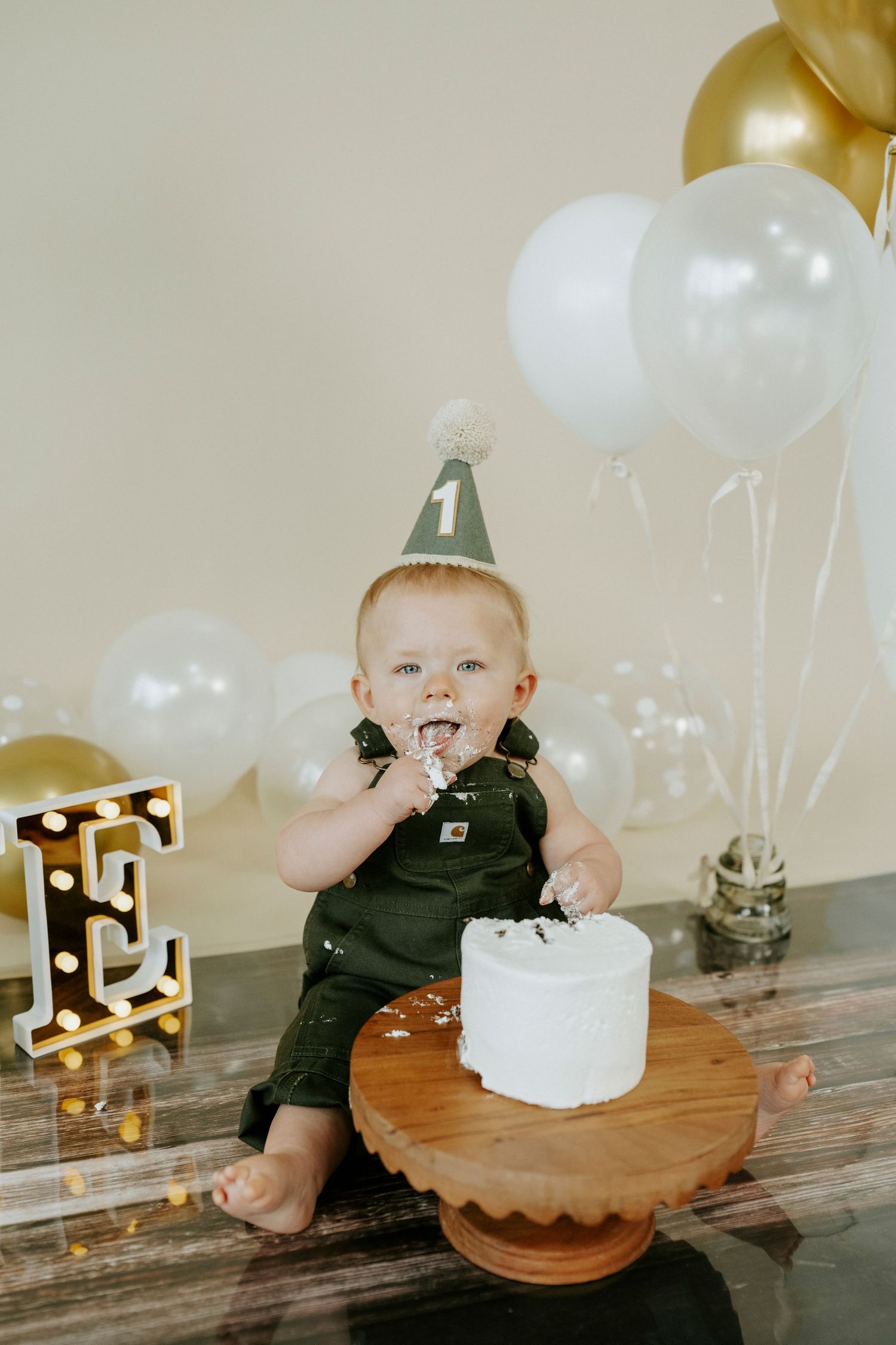 Cake smash photo session featuring white cake, gold decor, and white balloons for first birthday celebration.