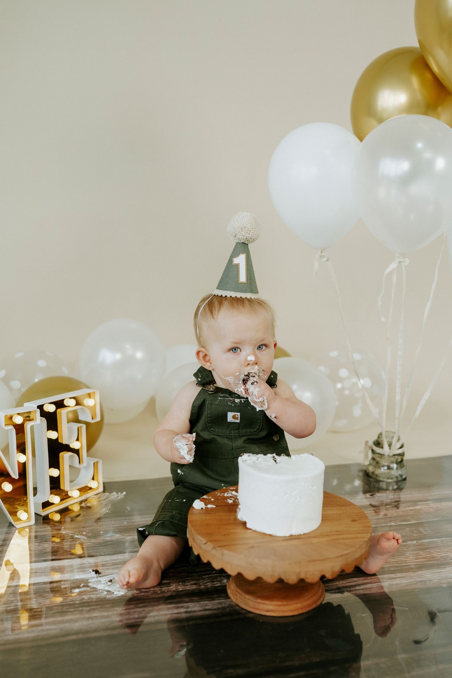 Cake smash photo session with white and gold balloons and illuminated number one sign.