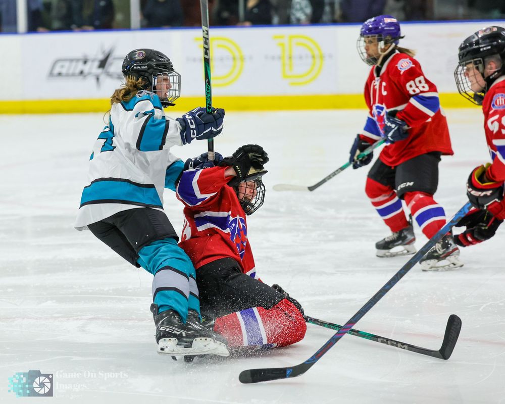Youth hockey players in teal and red jerseys battle for the puck during an intense game on the ice.