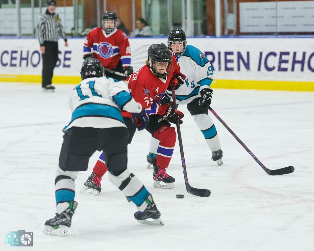 Youth hockey players in teal and red jerseys compete during an ice hockey game match.