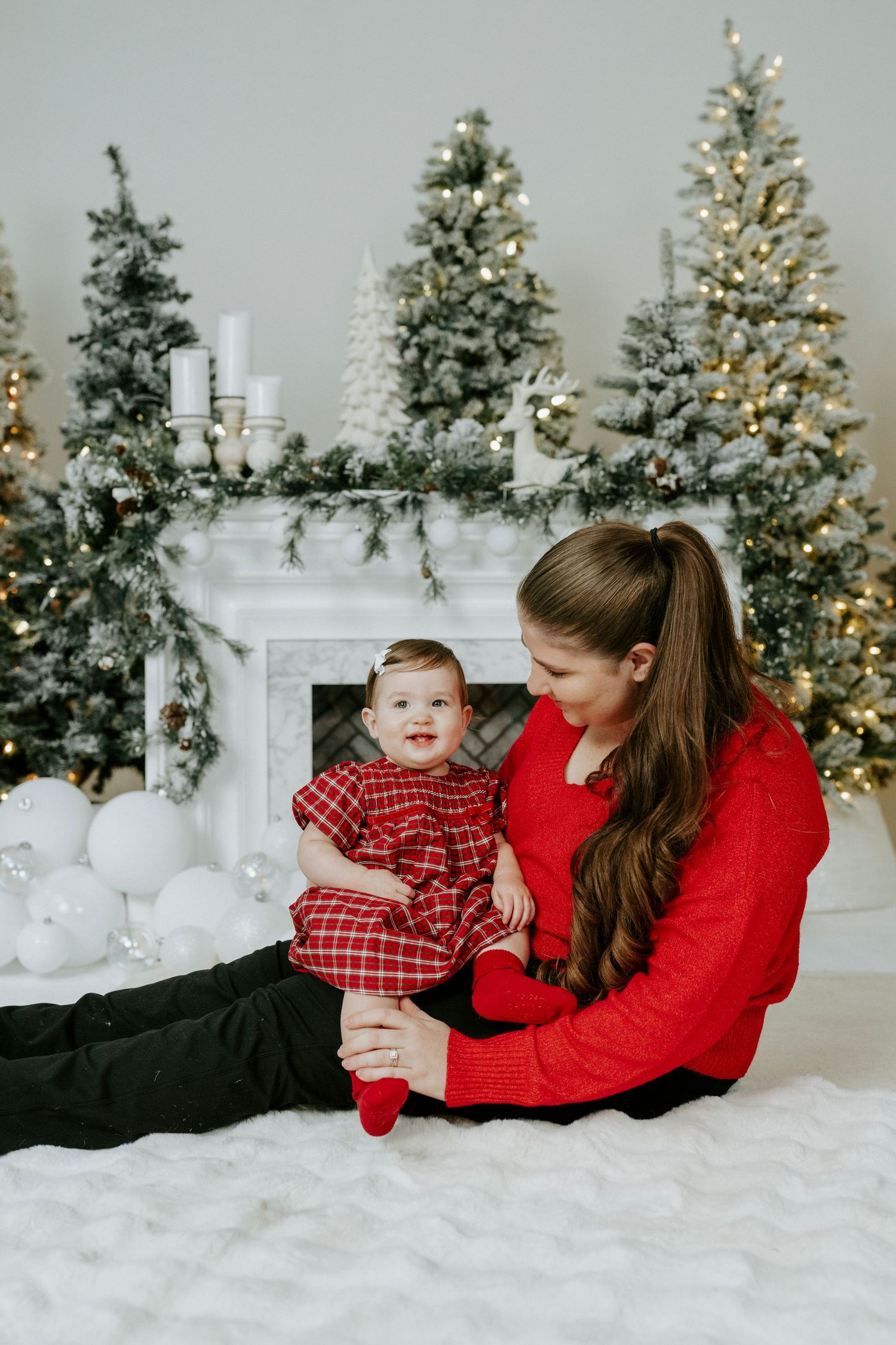 Mother and child in matching red outfits pose for Christmas photos with decorated trees and white fireplace in background.