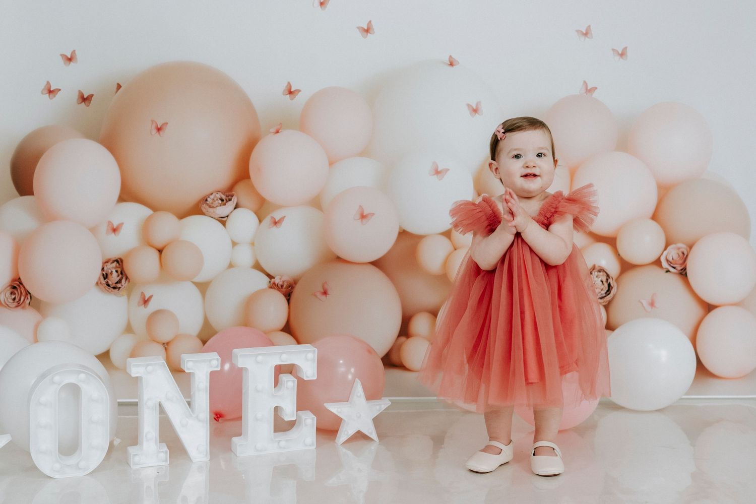 Baby in coral dress sits among pink and white balloon arch for first birthday celebration.