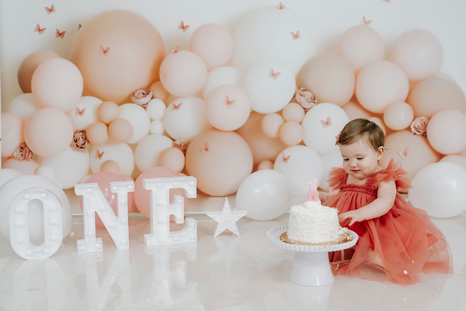 First birthday celebration setup with peach balloons, white ONE letters, and a cake against a soft pastel backdrop.