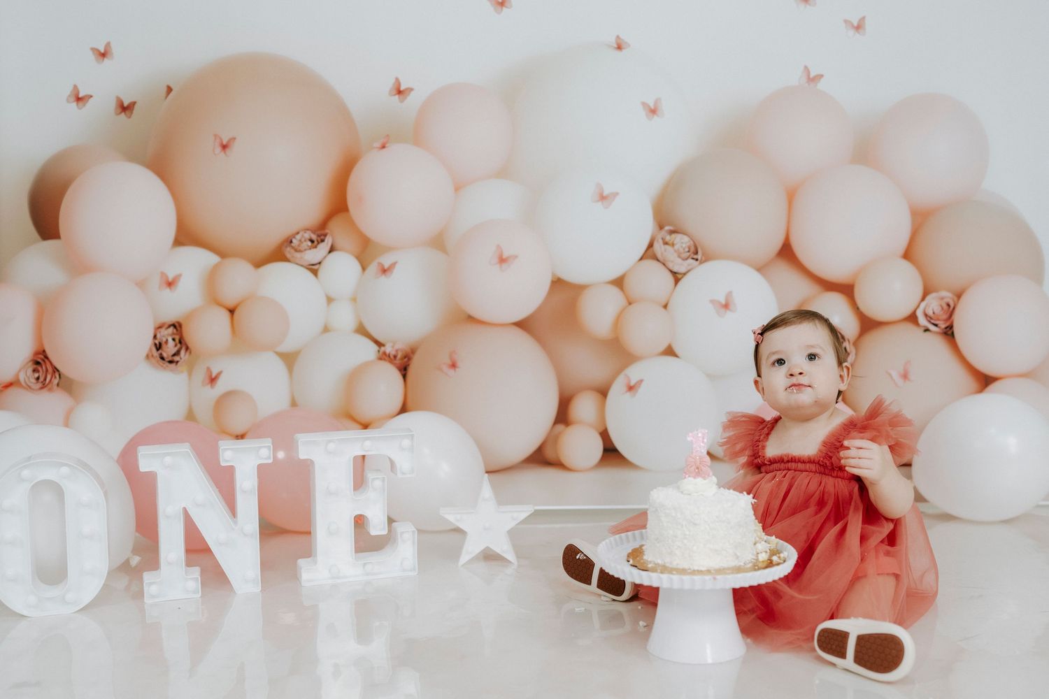 First birthday celebration setup with peach and white balloon arch, white 'ONE' letters, and cake on white floor.