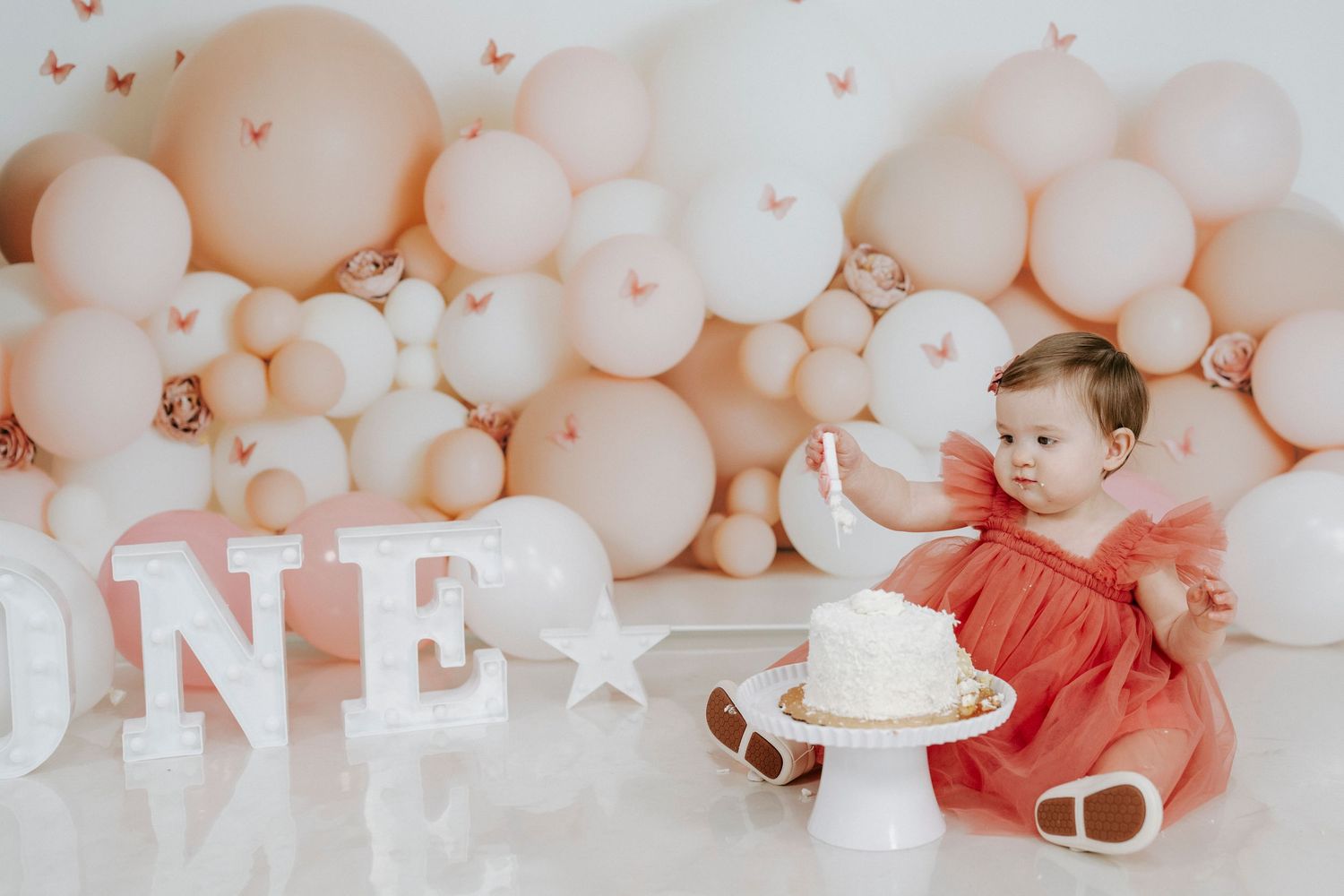 Baby in red dress sitting by peach balloon arch and white 'ONE' letters during first birthday photoshoot.