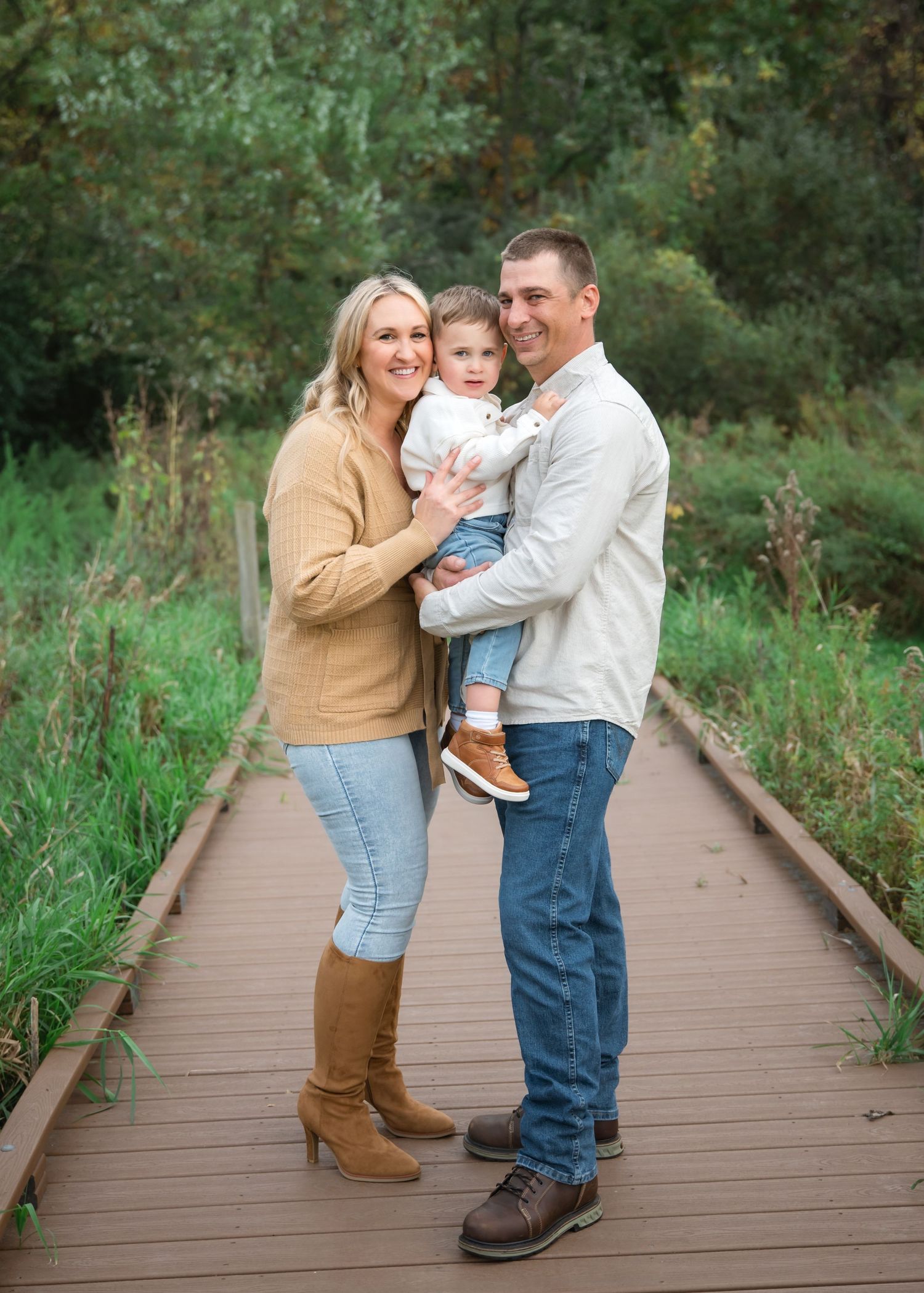 A family shares tender moments on a wooden boardwalk surrounded by lush greenery on a sunny day.