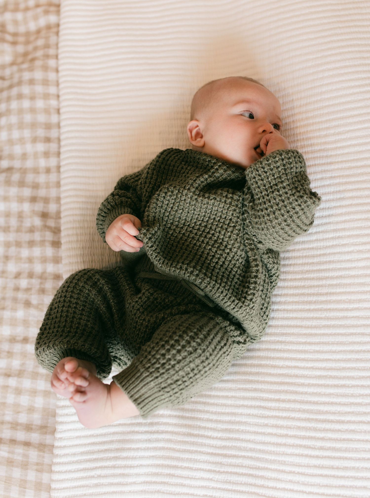 Baby in cozy olive green knit sweater lying on striped bedding in four sequential poses.