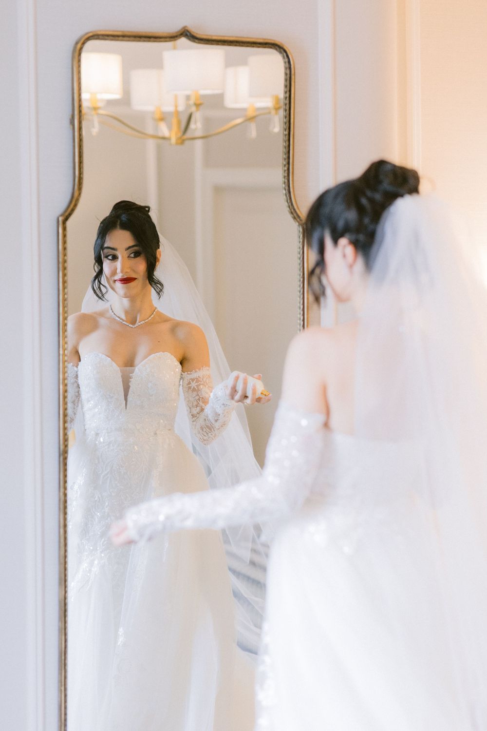 A bride in a white lace wedding dress with long sleeves looks at her reflection in an elegant gold-framed mirror.