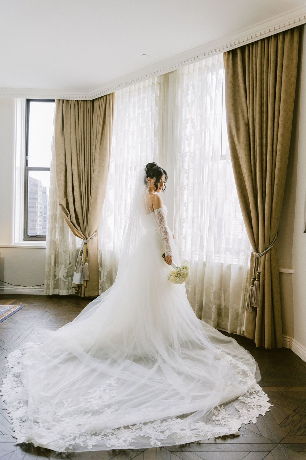 A flowing white wedding dress with an elegant long train displayed against white walls and sheer curtains in a bridal suite.