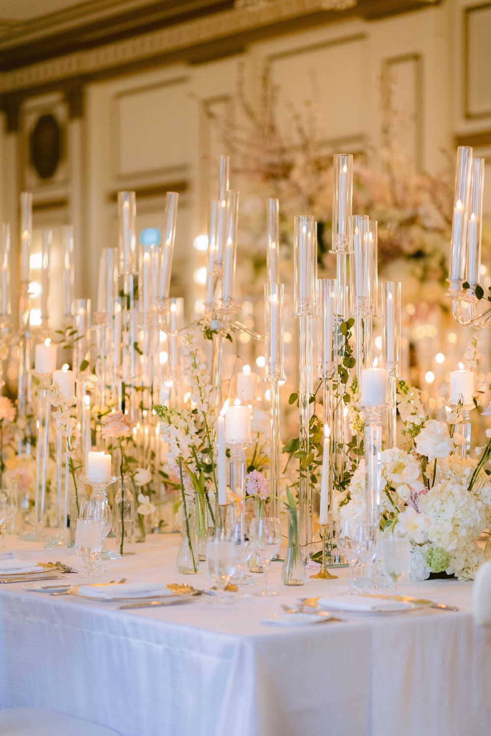 Long wedding reception tables decorated with white flowers and tall glass candle holders in an elegant setting.