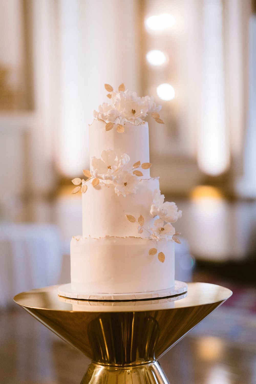 White wedding cake with delicate floral decorations sits atop a modern gold pedestal stand in elegant reception setting.