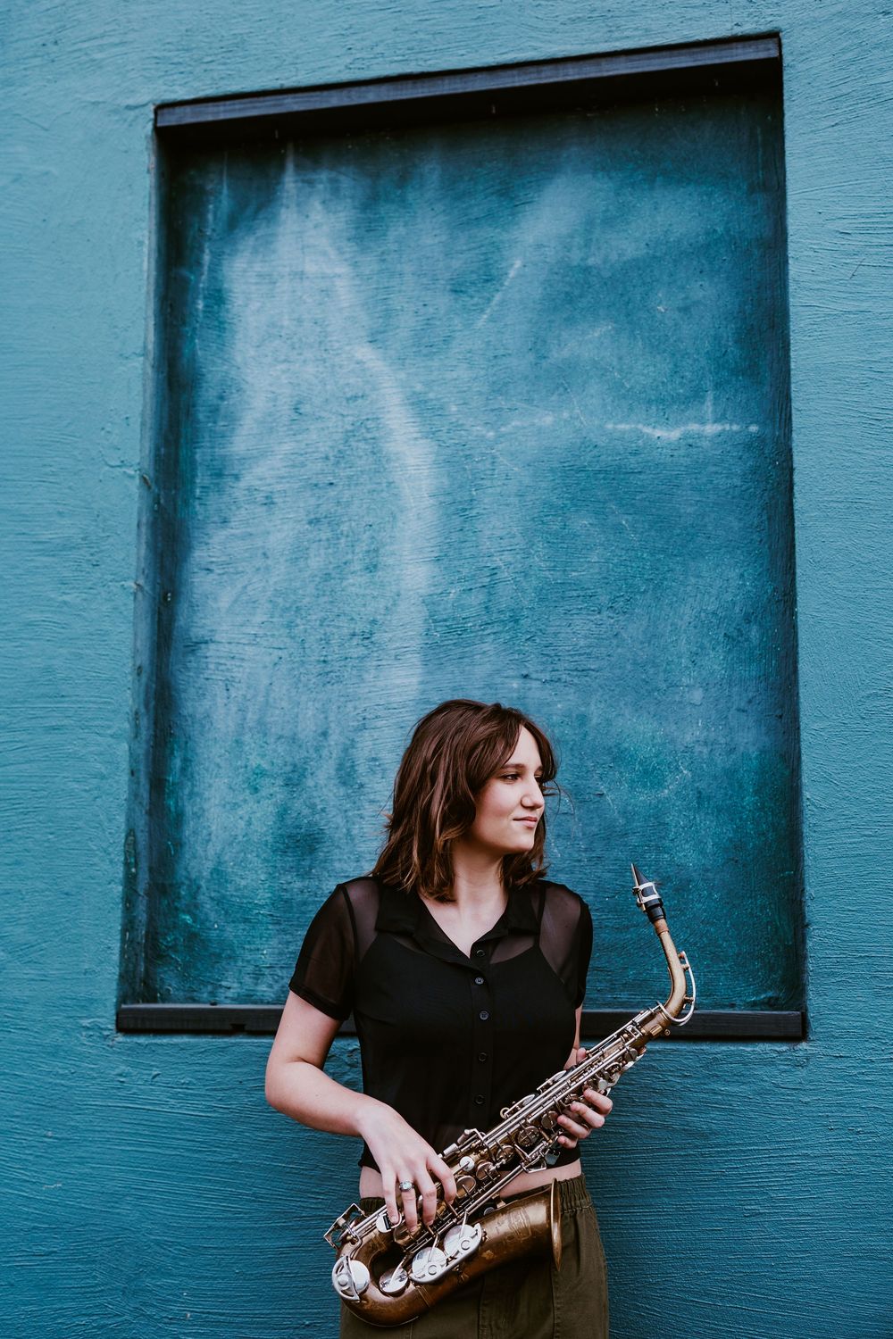 Saxophone player photographed against turquoise wall with window frames.