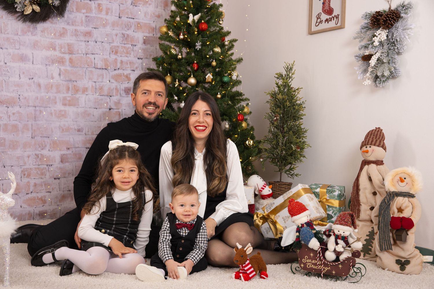 Family poses on floor with Christmas decorations and plush toys against brick wall with decorated tree.
