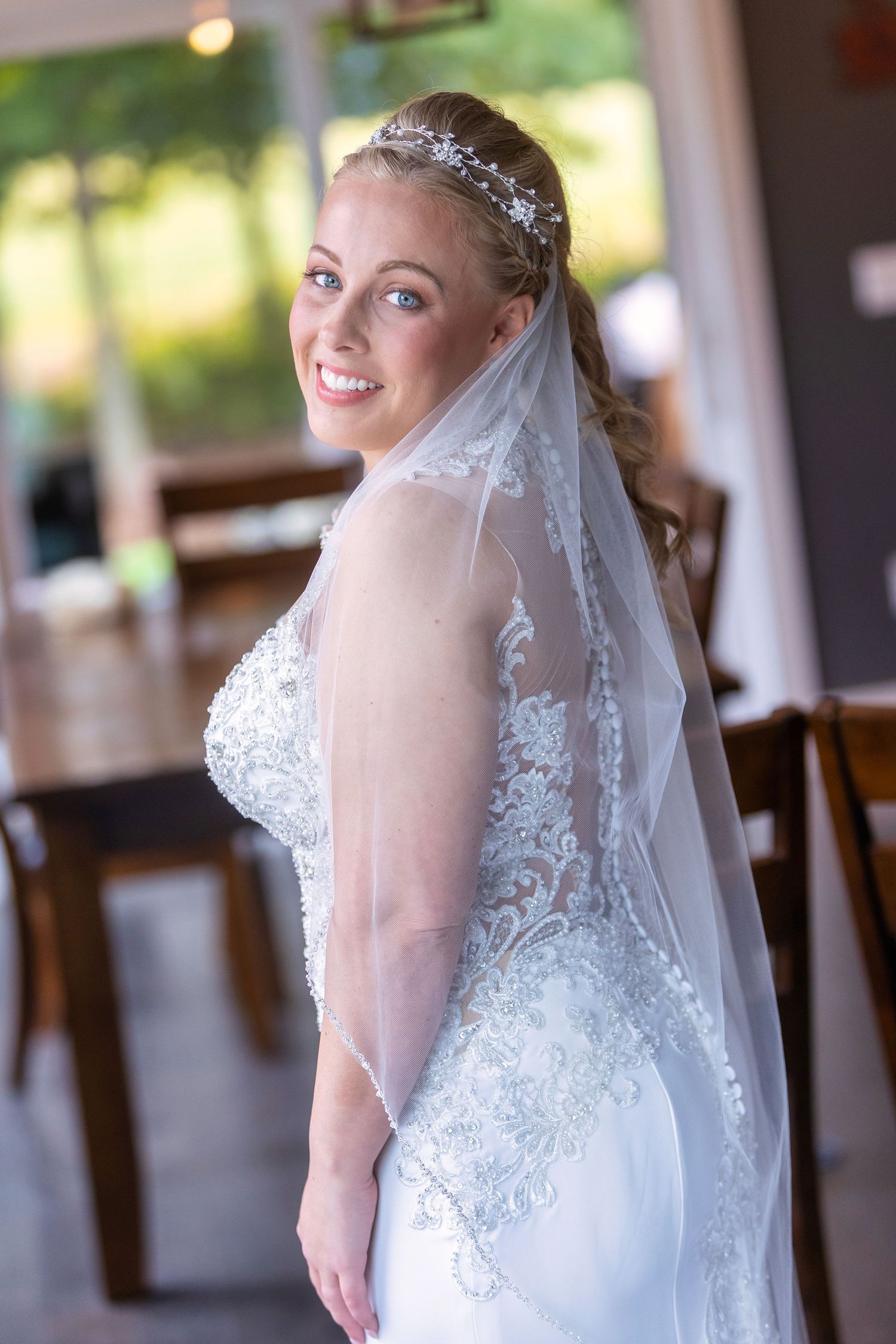 A bride in a white lace wedding dress with tiara and veil smiles while posing in an indoor venue.