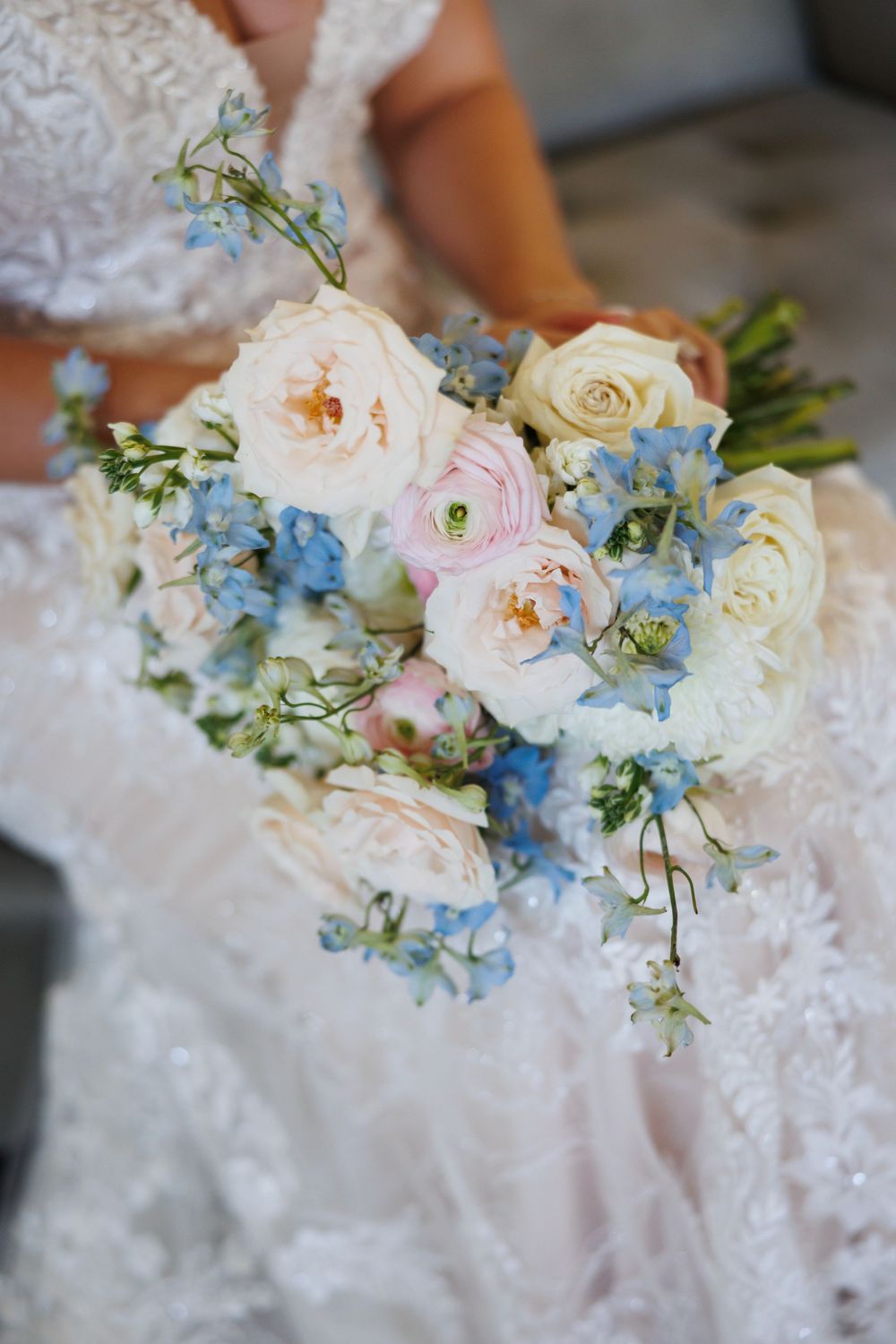 Delicate bridal bouquet featuring blush roses, white flowers, and blue delphiniums against white lace fabric.