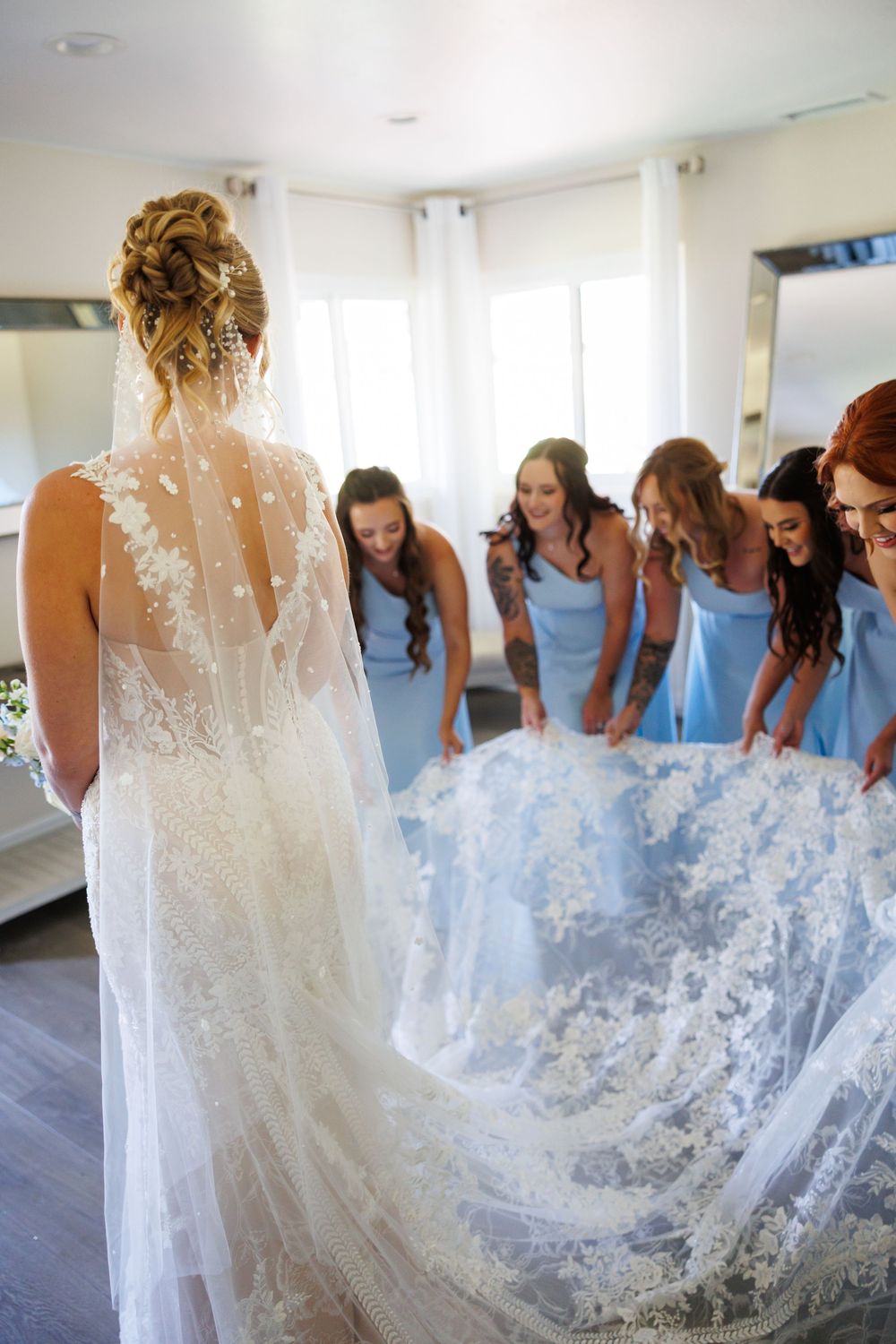 A bride in a flowing lace gown stands with bridesmaids wearing light blue dresses in a bright indoor setting.
