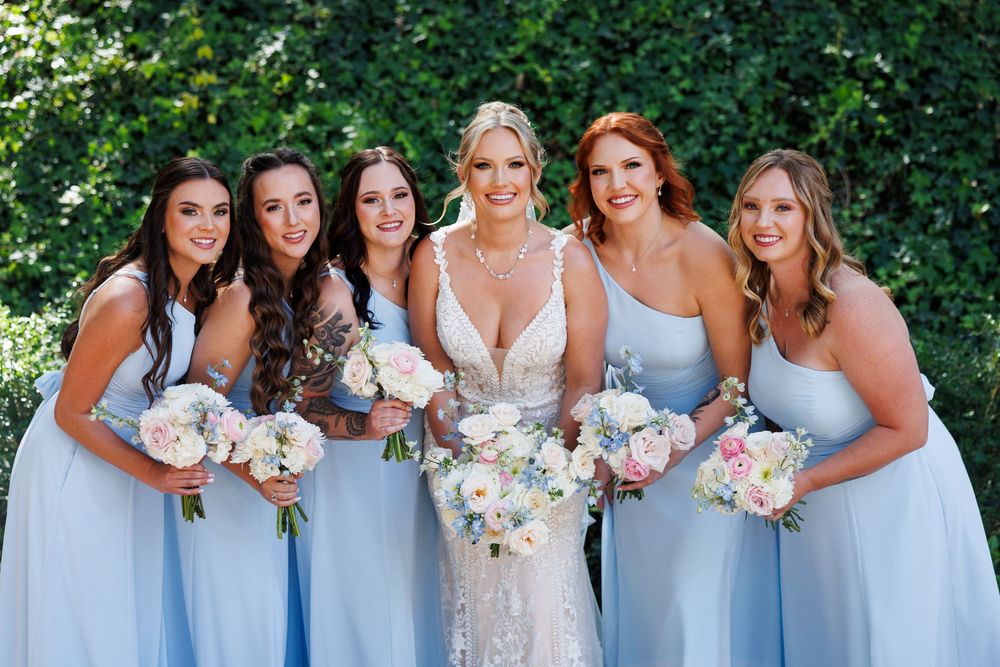 Wedding party in light blue bridesmaid dresses posing together with floral bouquets against green foliage.