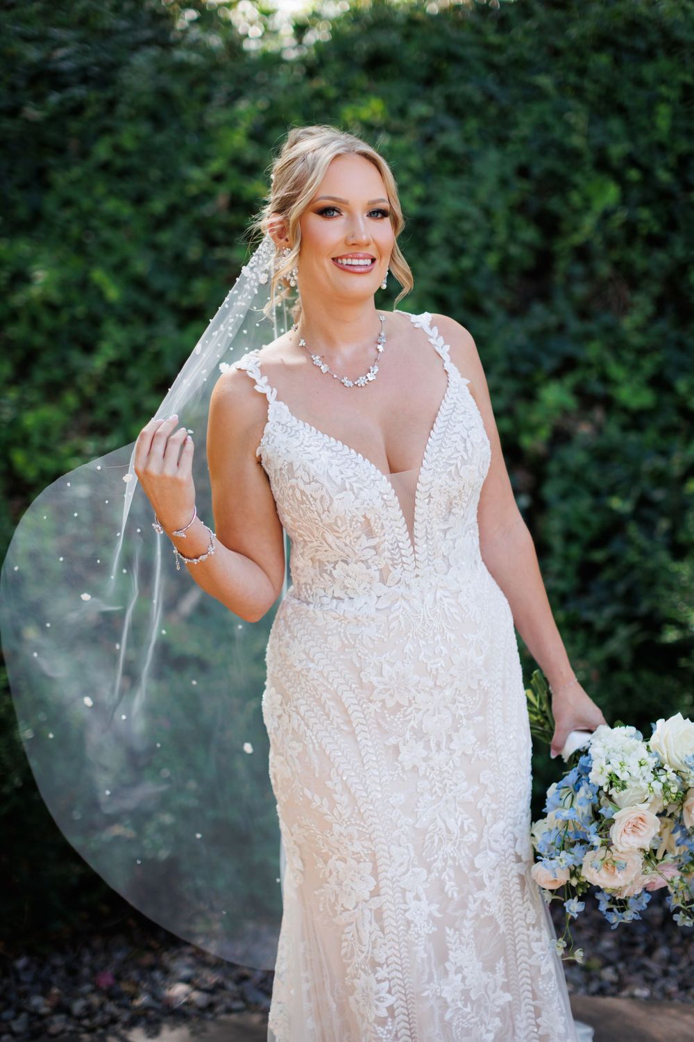 A bride in a sparkling white lace wedding gown with a cathedral veil poses against a green garden backdrop.