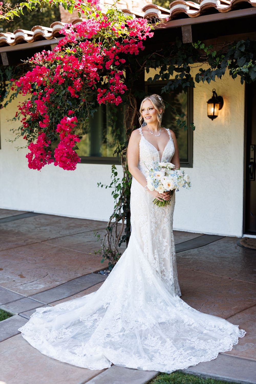 Elegant lace wedding dress with long train photographed against Spanish-style architecture with vibrant pink bougainvillea.